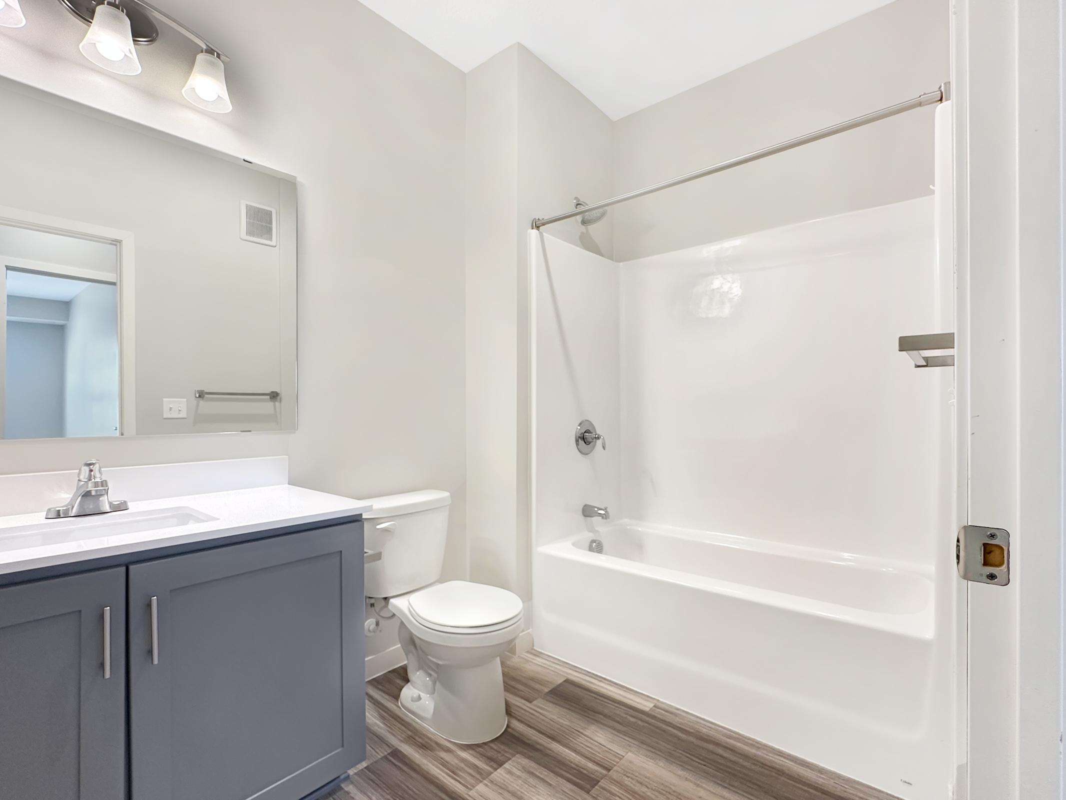 A modern bathroom featuring a white bathtub with an overhead shower, a large mirror above a dark blue vanity with a sink, a toilet, and neutral-toned walls. The floor has wood-like tiles for a contemporary look. Natural light may be present, enhancing the spacious feel.