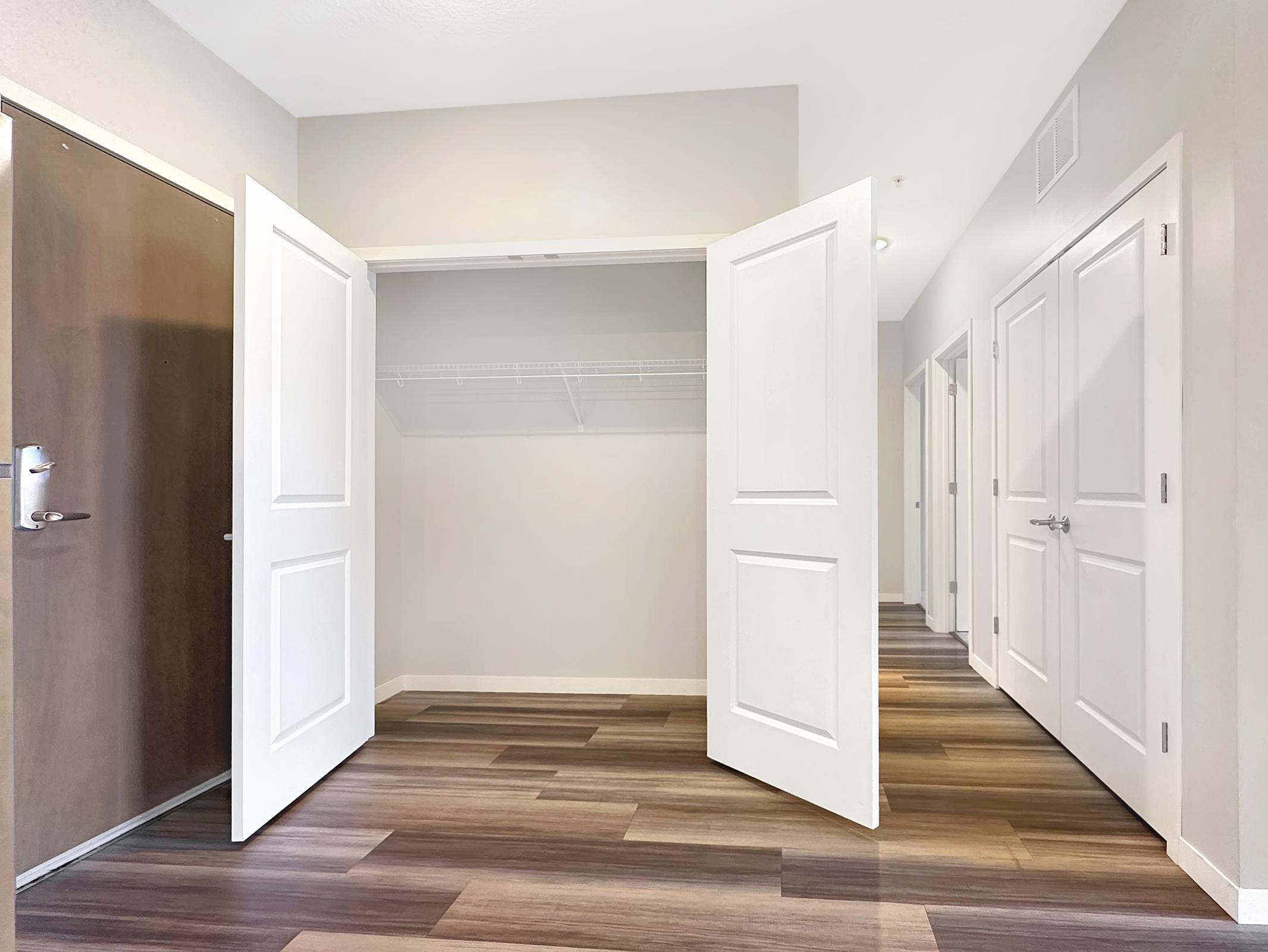 A spacious hallway with open white closet doors on the left, displaying a wire shelving unit. The floor features modern wood-like planks, and walls are painted in neutral tones. Additional doors lead to other rooms in the background, creating a bright and airy atmosphere.