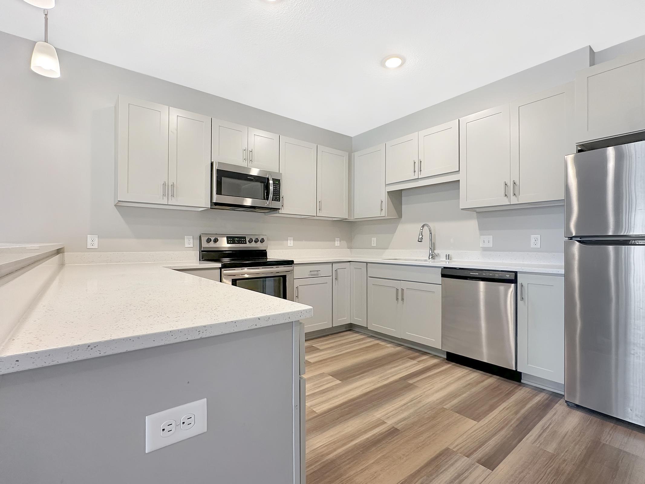 Modern kitchen featuring light gray cabinets, stainless steel appliances including a microwave and refrigerator, a sleek countertop, and open layout with wood-like flooring. The space is well-lit with natural light and recessed lighting.