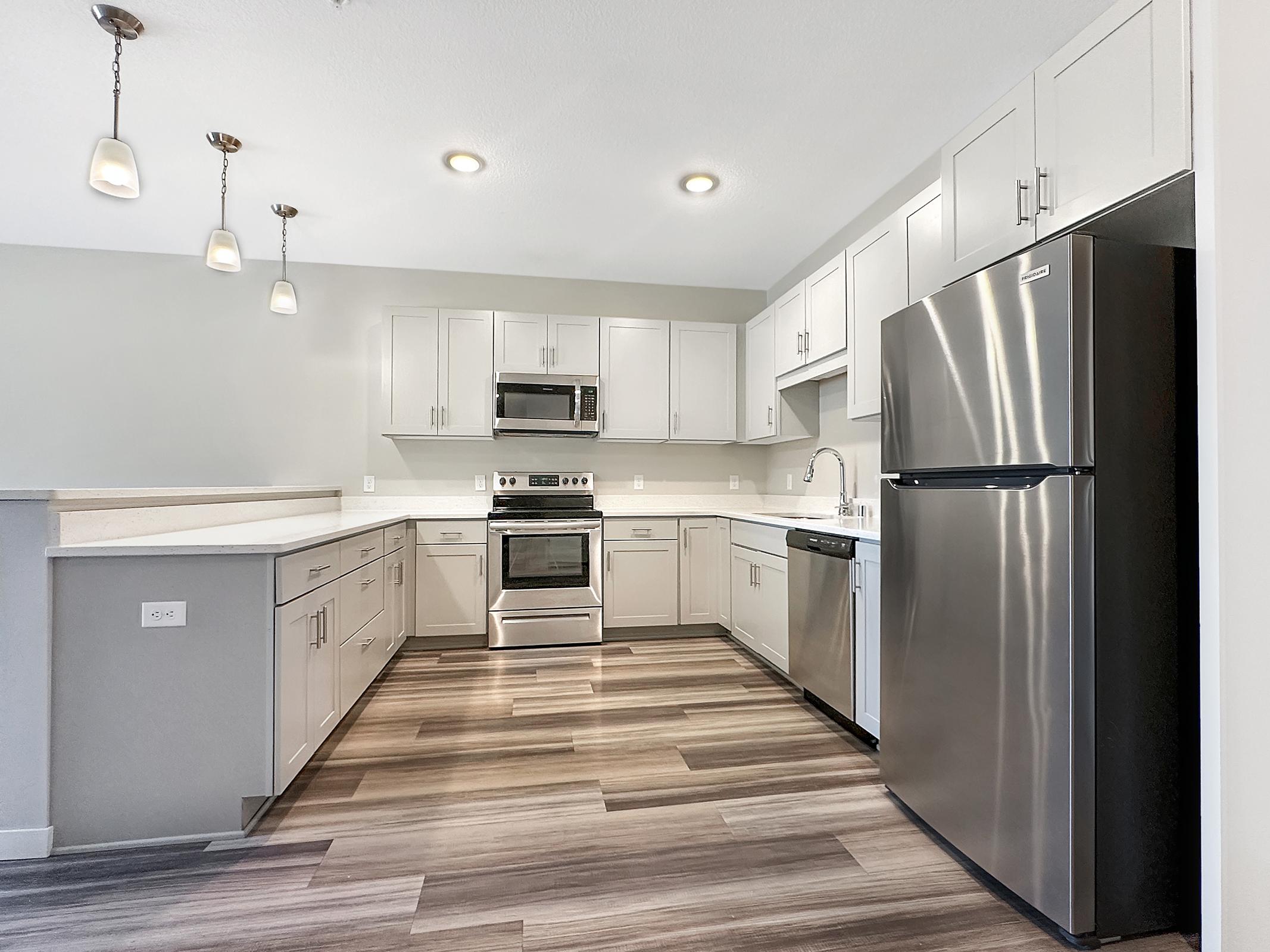 Modern kitchen featuring gray cabinets, stainless steel appliances, an undermount sink, and quartz countertops. The space is illuminated by pendant lighting and has a light wood-like laminate floor. A sleek refrigerator and a built-in microwave add to the contemporary design.