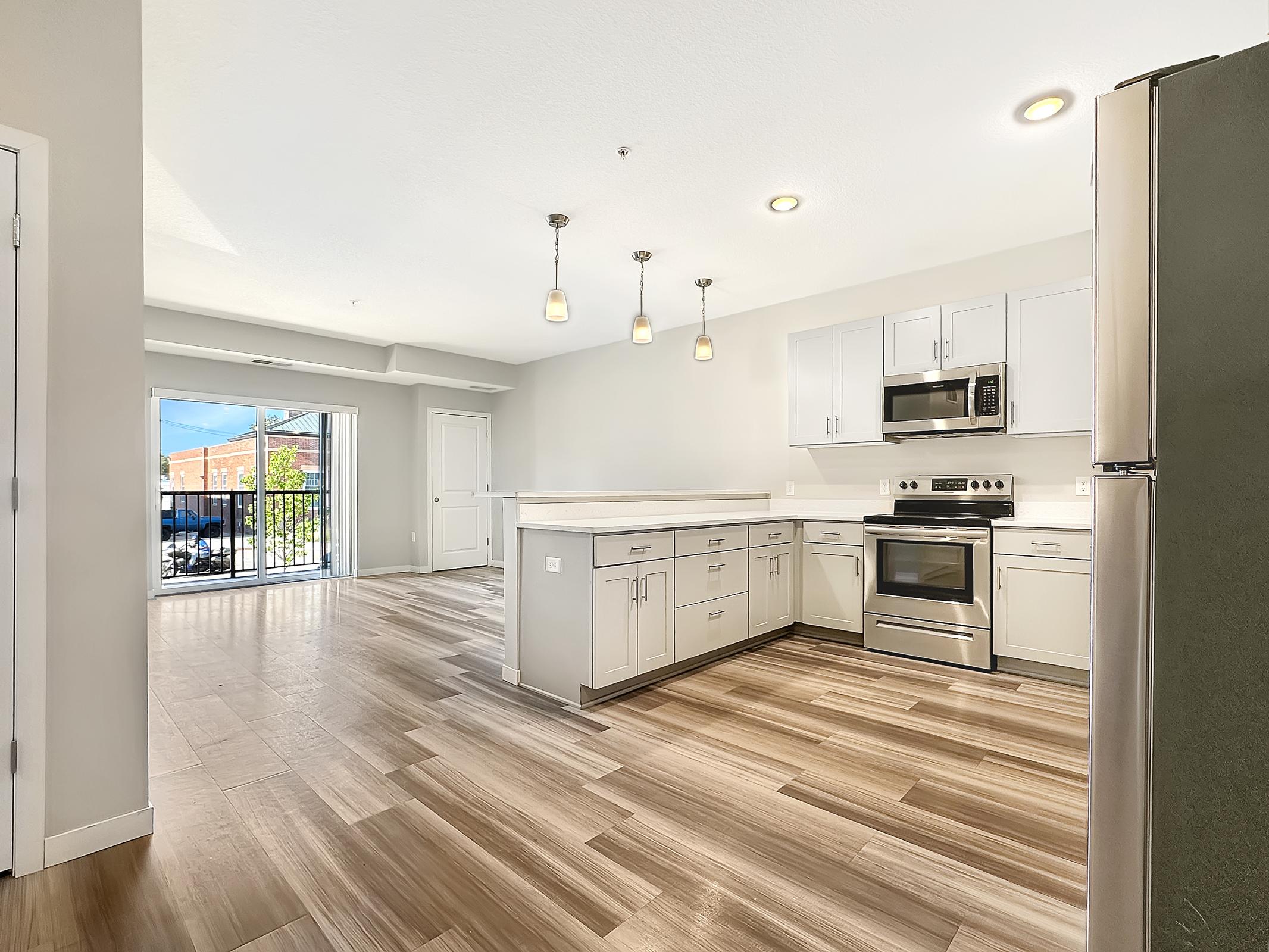 Modern kitchen featuring stainless steel appliances, including a microwave and oven. It has light gray cabinetry, a large island with a countertop, and pendant lighting. The hardwood floor adds warmth, and there's a sliding door leading to an outdoor area, providing natural light and an open feel.