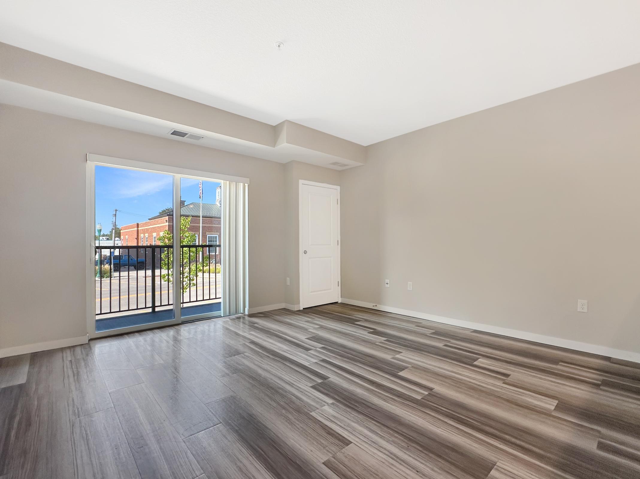 Spacious interior of an apartment featuring light-colored walls and modern wood-style flooring. Large sliding glass door opens to a balcony, allowing natural light to fill the room. Neutral tones create a welcoming atmosphere, and the layout is open and versatile for furnishings.