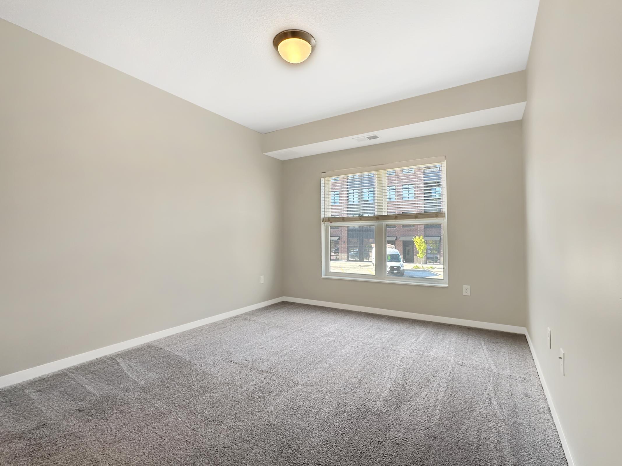Empty room with light gray walls and carpeted flooring. A single ceiling light fixture illuminates the space. Large window with blinds allows natural light to enter, revealing a view of a building outside. The room has a minimalist feel, suitable for various layout options.