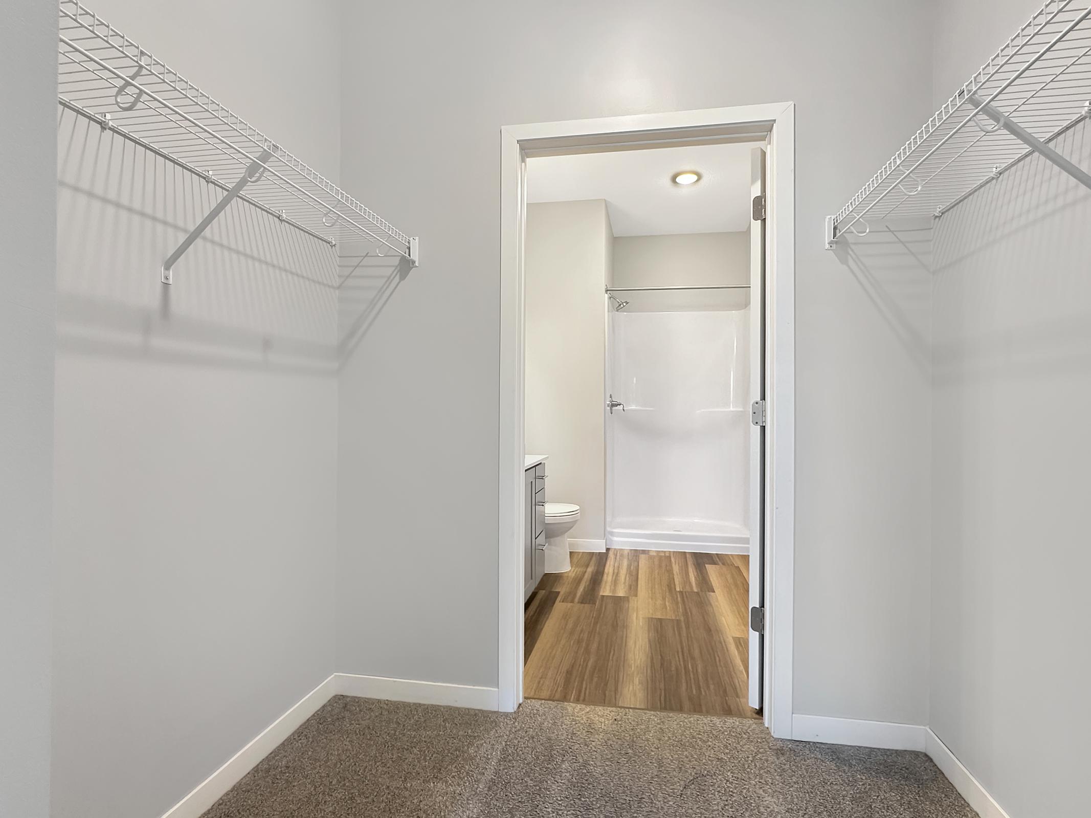 A spacious closet with wire shelving on both sides, leading to a bathroom area with a shower. The walls are painted a light color, and the floor features wood-like tiles. The closet is well-lit, creating a bright and open feel.