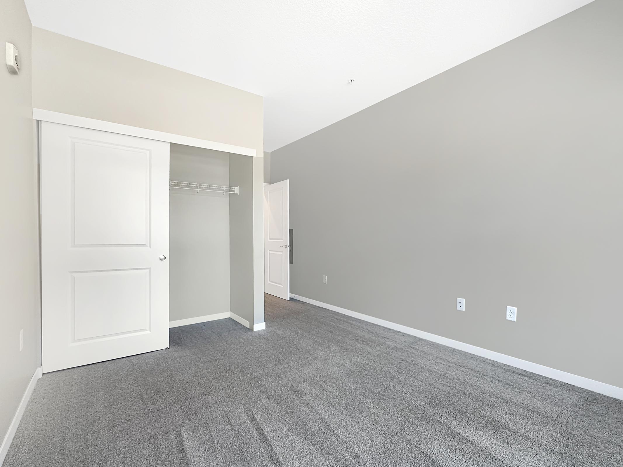 A spacious, minimally decorated room featuring gray walls and carpet. There's a closet with a white door on the left, and the room has a clean, modern appearance with ample natural light. The overall design is simple and inviting, ideal for personal customization.