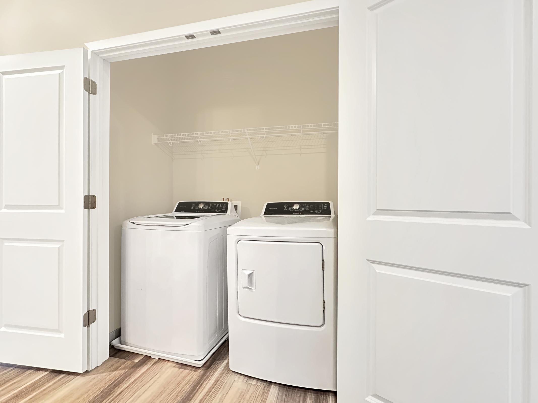 A laundry room with a white washing machine and a dryer side by side. The appliances are set against a light-colored wall, with a shelf above them for storage. The space is partially enclosed by two doors, and the flooring is a wooden-style laminate.
