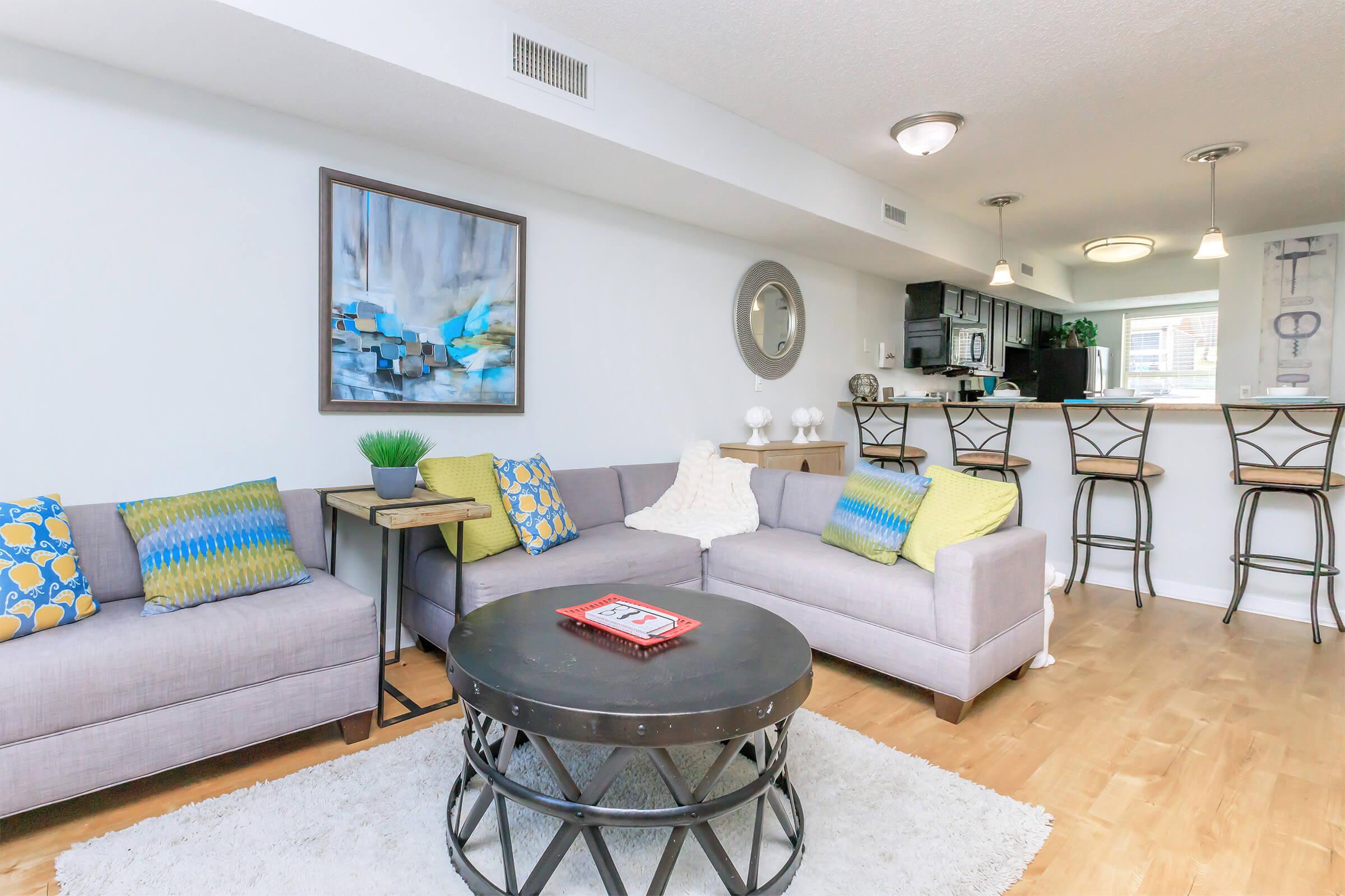 Cozy living room featuring a gray sectional sofa with colorful throw pillows, a round coffee table, and a vibrant piece of artwork on the wall. A small side table with a potted plant adds a touch of greenery. The open layout leads to a kitchen with bar seating in the background, creating a bright and inviting atmosphere.