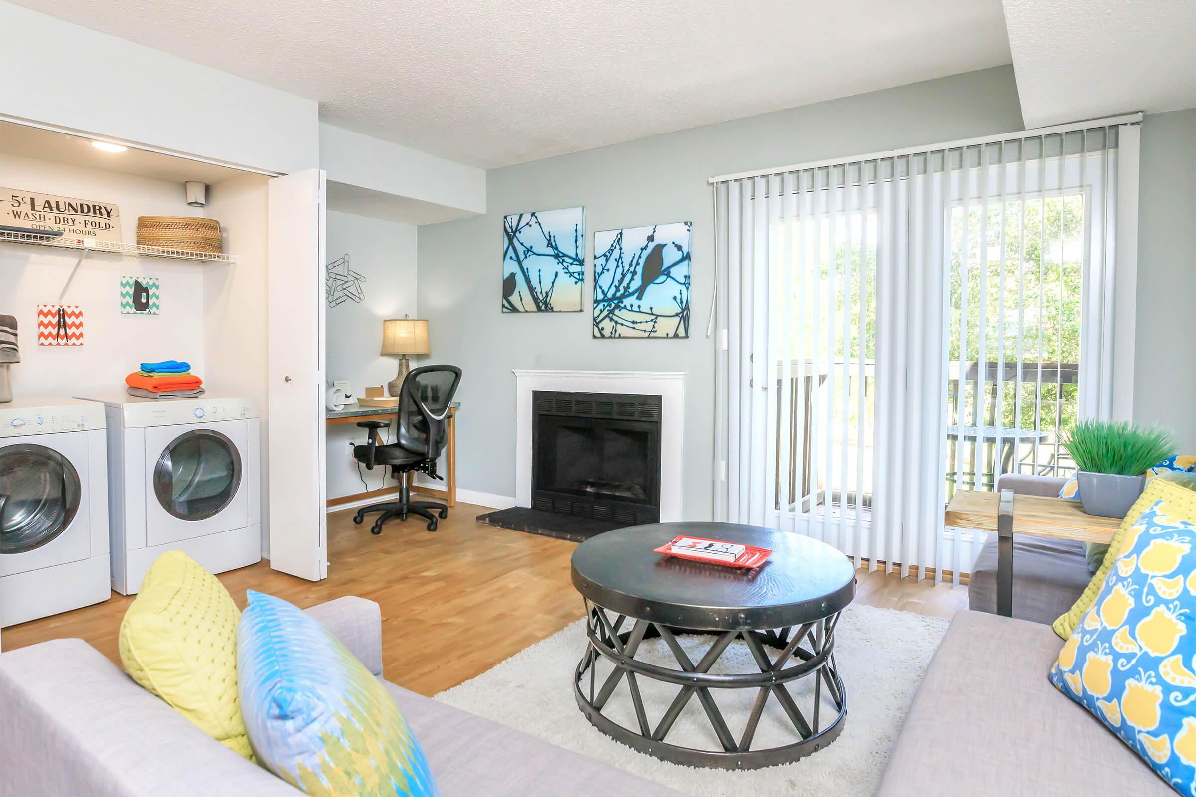 A cozy living room featuring a light gray wall, a comfortable sofa with blue and yellow cushions, a round coffee table, and a modern aesthetic. There's a small workspace with a desk and lamp, a laundry area with stacked washing machines, and a sliding door leading to a balcony.