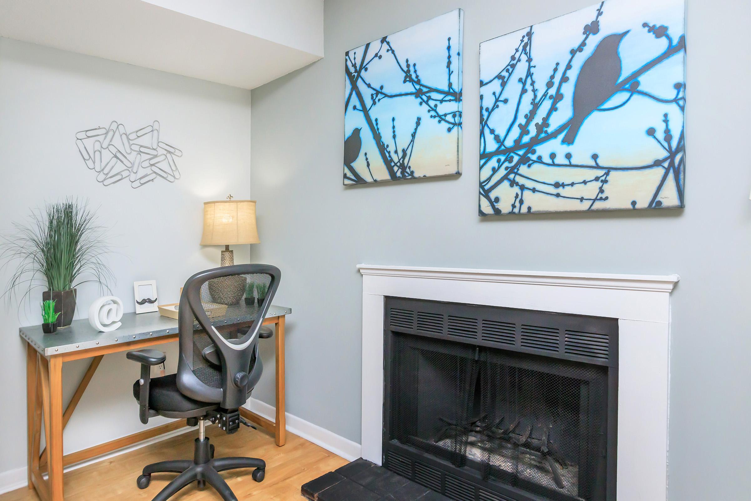 A cozy home office setup featuring a modern desk with a black chair, a lamp, and decorative elements. The wall is adorned with two artistic bird-themed paintings in blue tones, and there's a small plant on the desk. A fireplace with a black surround adds warmth to the space.
