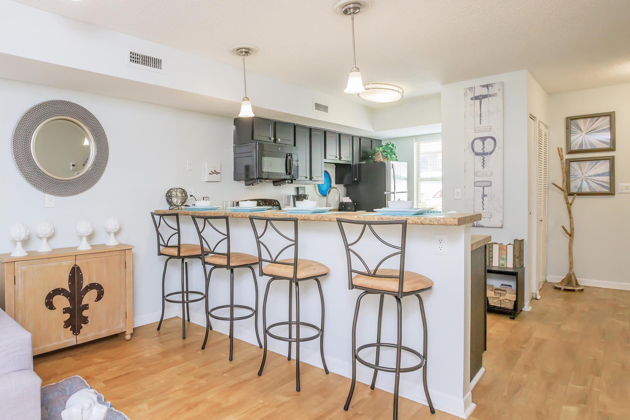A modern open-concept kitchen and living area featuring a bar with four stools, dark cabinets, and a light countertop. A decorative cabinet with a fleur-de-lis design sits adjacent, and decorative wall art adorns the space, creating a stylish and inviting atmosphere.