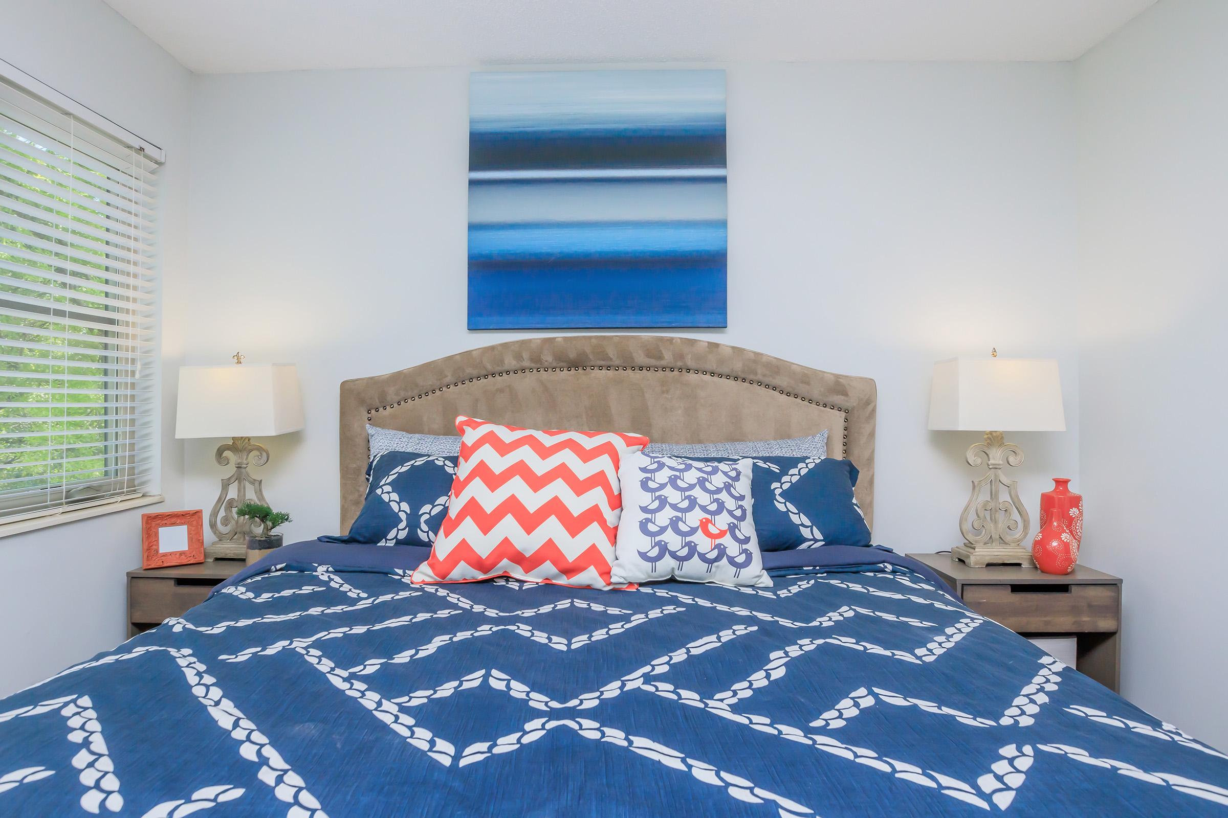 A cozy bedroom featuring a large bed with a blue and white patterned duvet and colorful decorative pillows. Two bedside lamps illuminate the space, and a piece of abstract artwork in shades of blue hangs above the headboard. A window with blinds allows natural light to enter the room.
