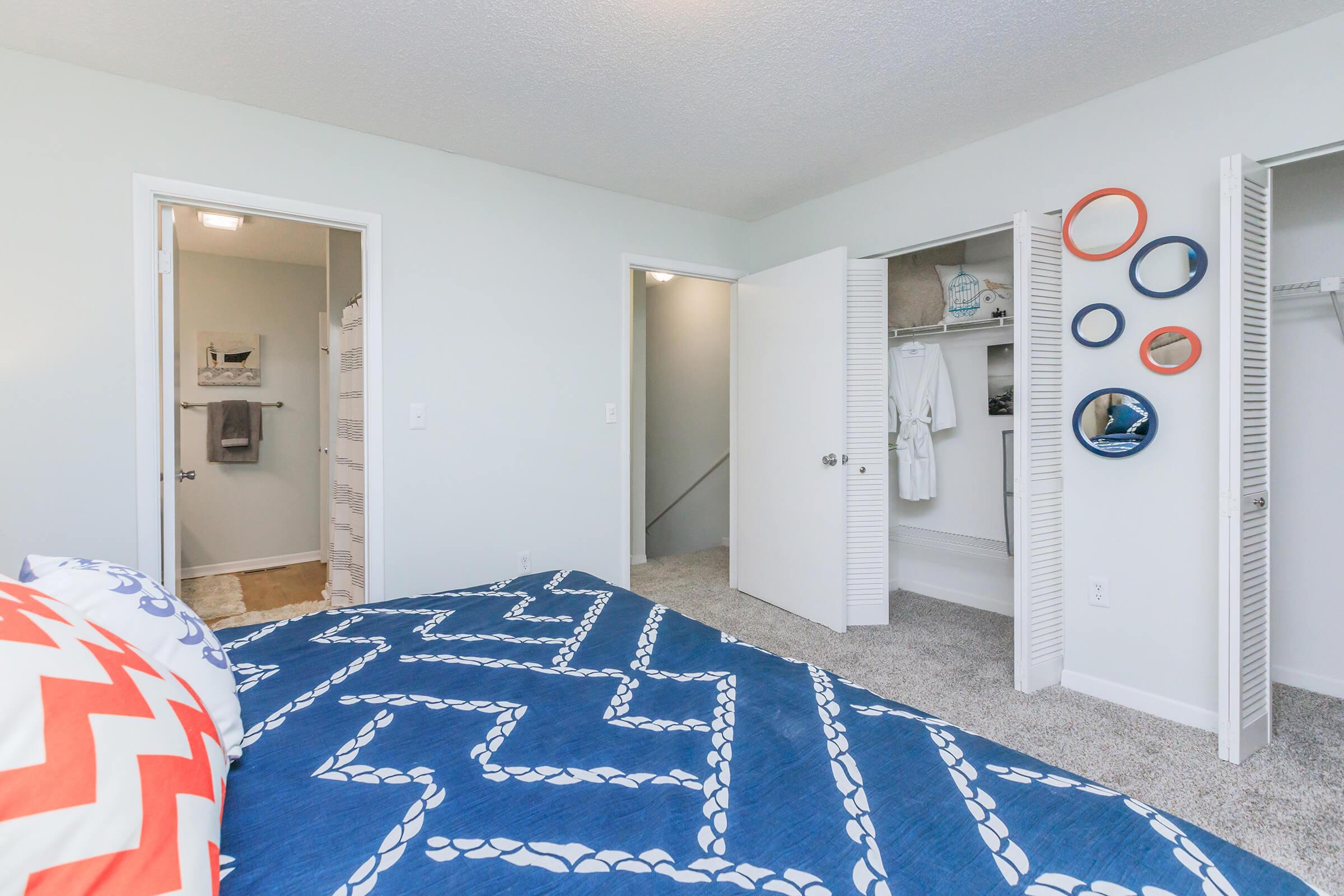 A well-lit bedroom featuring a blue and white chevron patterned comforter. There are two open closet doors revealing shelves and storage. The walls are painted a soft light color, providing a clean and modern look. A bathroom door is visible on the left, enhancing the room's functionality.