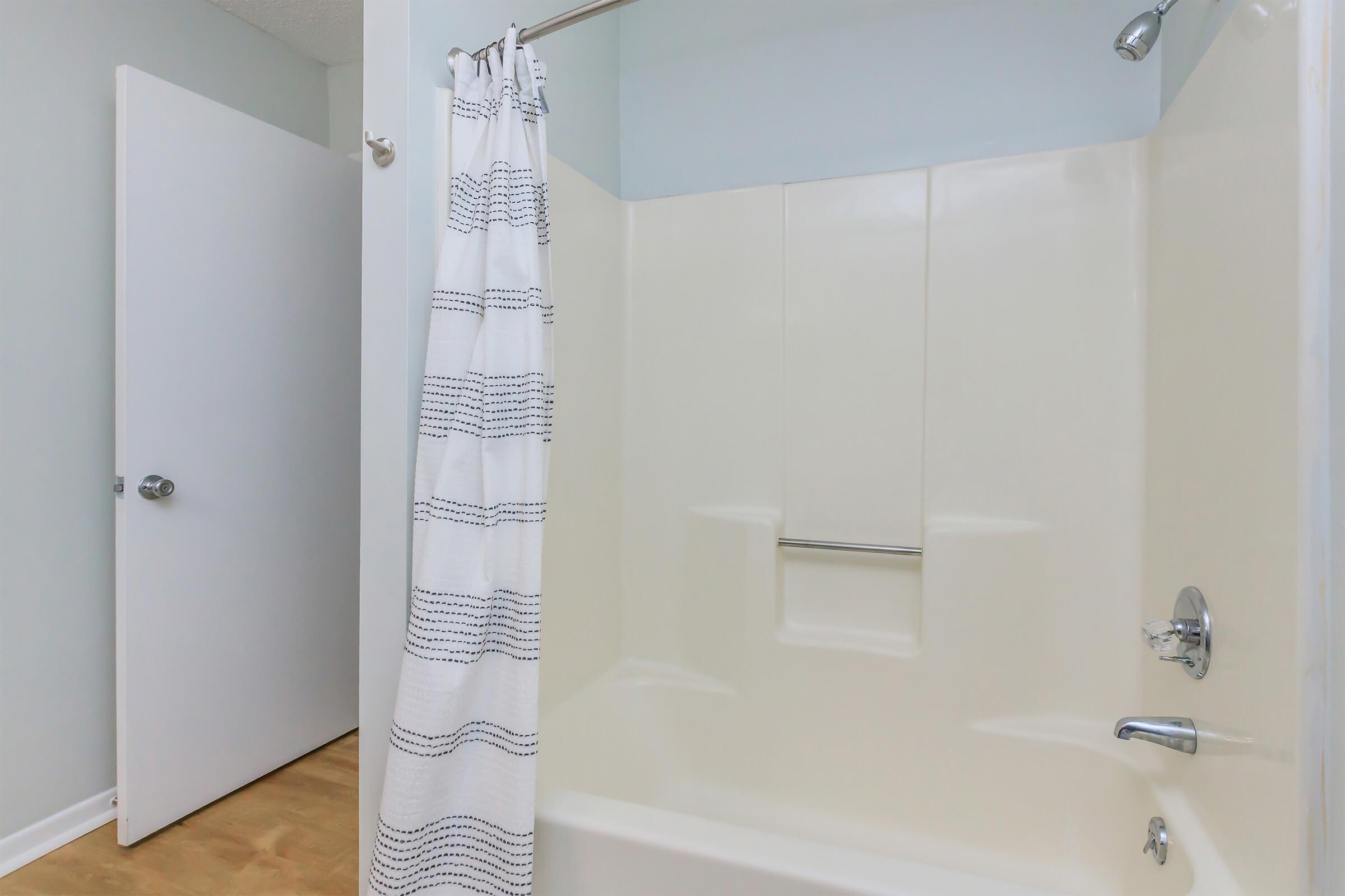 A clean, well-lit bathroom featuring a shower with a white curtain, a light-colored shower enclosure, and a door leading to another room. The floor is wooden, and the walls are painted in a soft hue, providing a serene and tidy atmosphere.