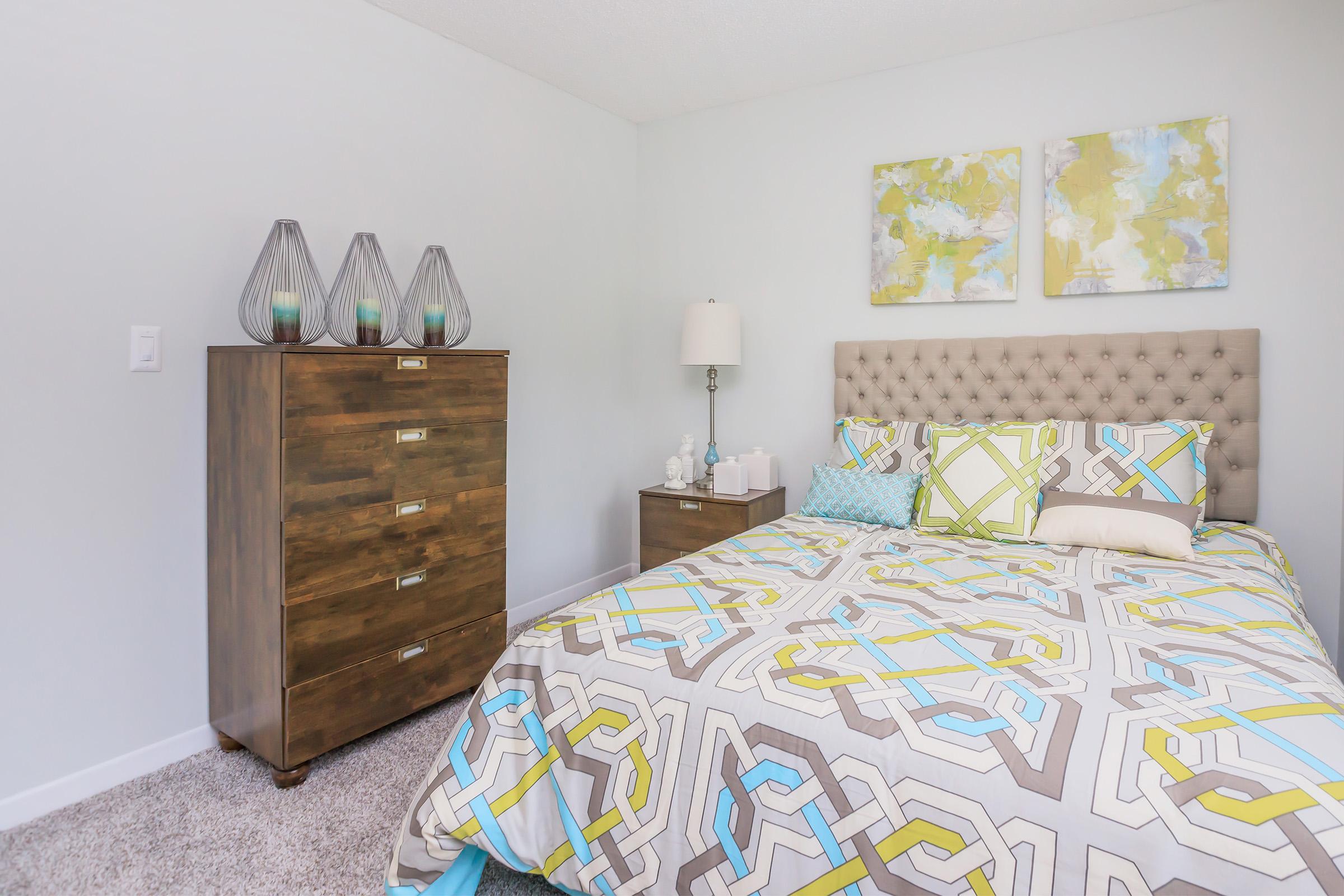 A well-decorated bedroom featuring a plush, tufted headboard bed with a colorful geometric patterned comforter. There is a wooden dresser with drawers, a bedside table with a lamp, and decorative vases on the dresser. The walls are painted light colors, and there are abstract artworks above the bed.