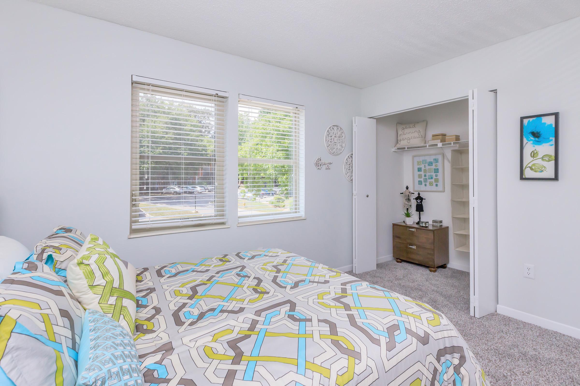 A bright and airy bedroom with a patterned comforter in shades of blue and yellow. The room features two windows allowing natural light, a nightstand with decor, and a closet with open shelving. The walls are painted light gray, enhancing the spacious feel of the room.
