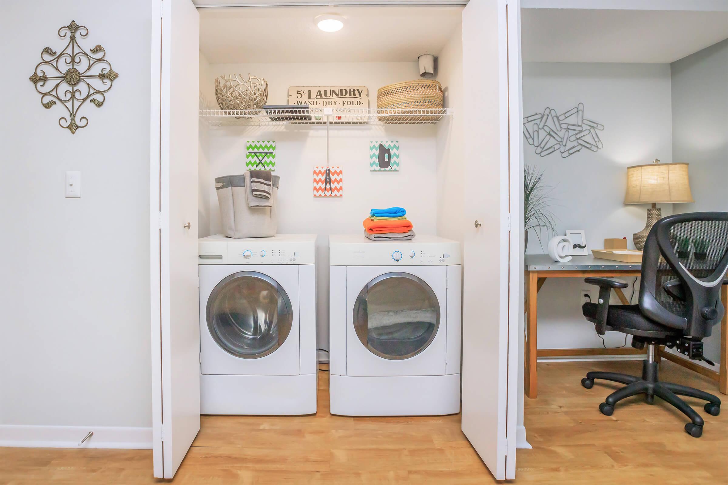 A laundry closet with two white washing machines side by side. Above, there are shelves holding a decorative basket and colored towels. Hanging from the shelves are three colorful towels. To the side, there is a desk with an office chair and a small decorative item. The walls are light gray with minimal decor.