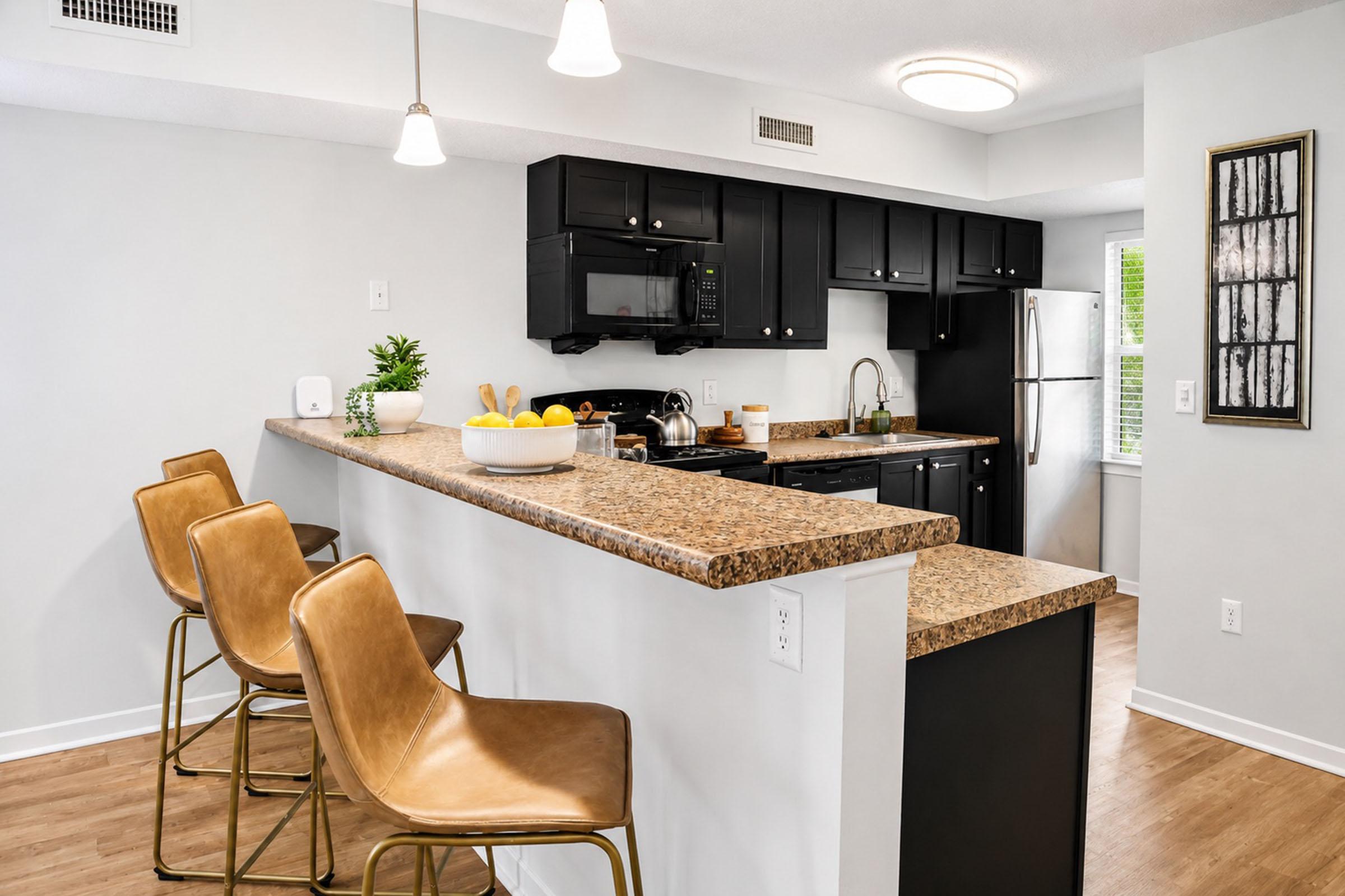 Modern kitchen with black cabinets and granite countertops. A bar area with three stylish, tan high stools is visible. Above, pendant lights illuminate the space. The kitchen features a stove, microwave, and a bowl of fresh fruit on the counter, with a window allowing natural light to enter.