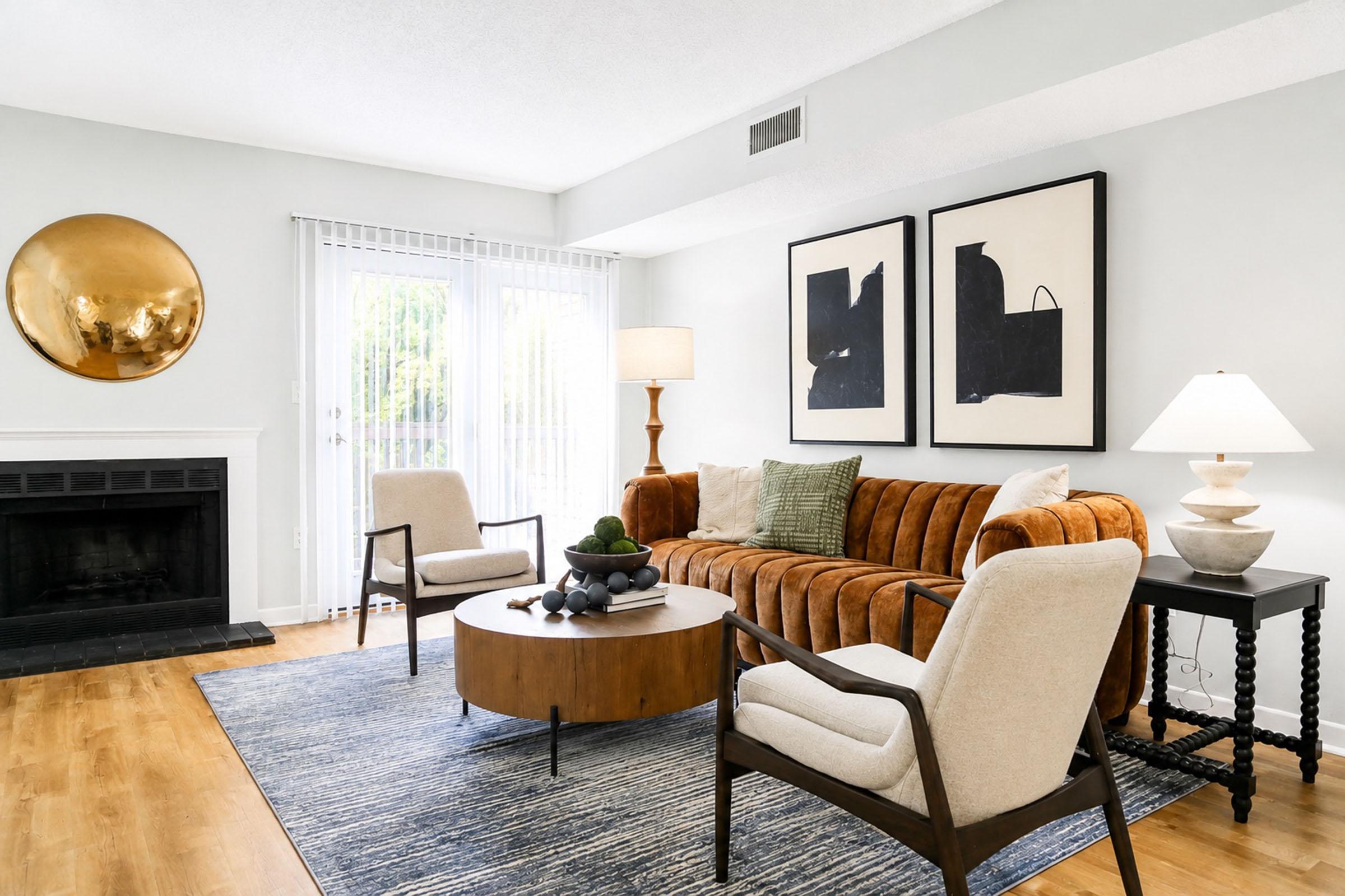 A modern living room featuring a cozy brown sofa, two light-colored armchairs, and a round wooden coffee table. There is a large circular mirror on the wall, abstract art pieces framed above the sofa, and a floor lamp. A fireplace is visible, along with sliding glass doors that open to an outdoor space.