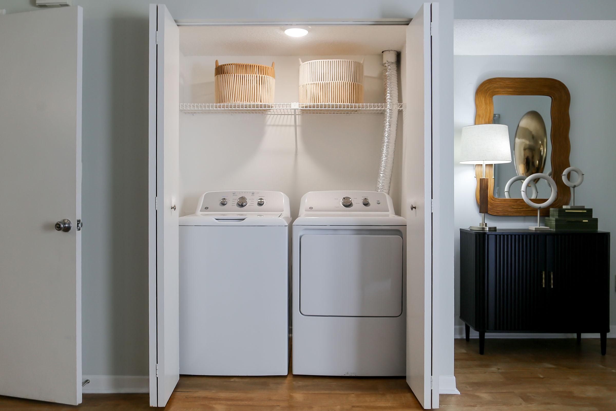 A laundry closet featuring a stacked washing machine and dryer. Above them are two woven baskets on a shelf. The closet has white doors that are open, revealing the appliances. A nearby black cabinet and a decorative mirror can be seen in the background, along with a lamp providing soft lighting.