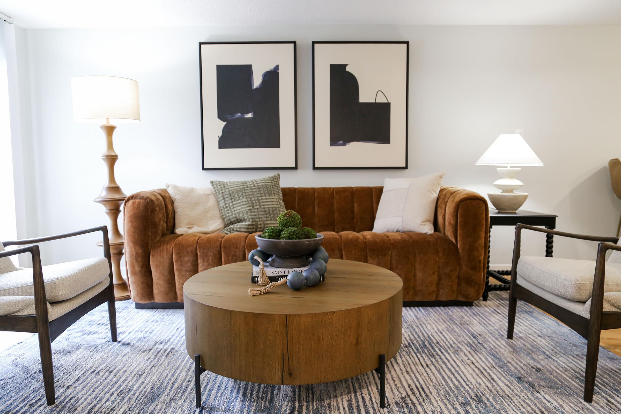 A cozy living room featuring a brown velvet sofa with decorative pillows, a round wooden coffee table, and two armchairs. The space is adorned with abstract black and white wall art and is well-lit by a floor lamp and a table lamp, creating a warm and inviting atmosphere.