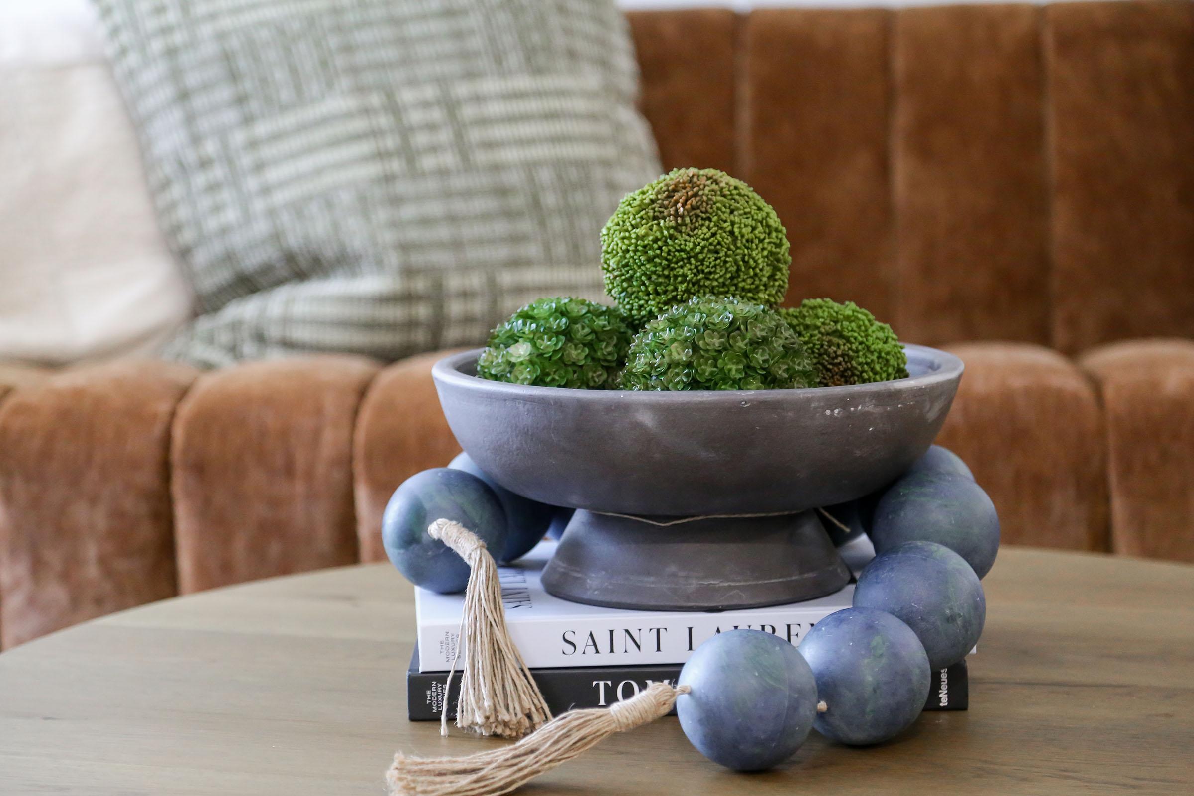A decorative bowl filled with green moss balls sits atop a stack of books on a wooden table. The background features a cozy brown couch with textured pillows, creating a warm and inviting living space atmosphere.