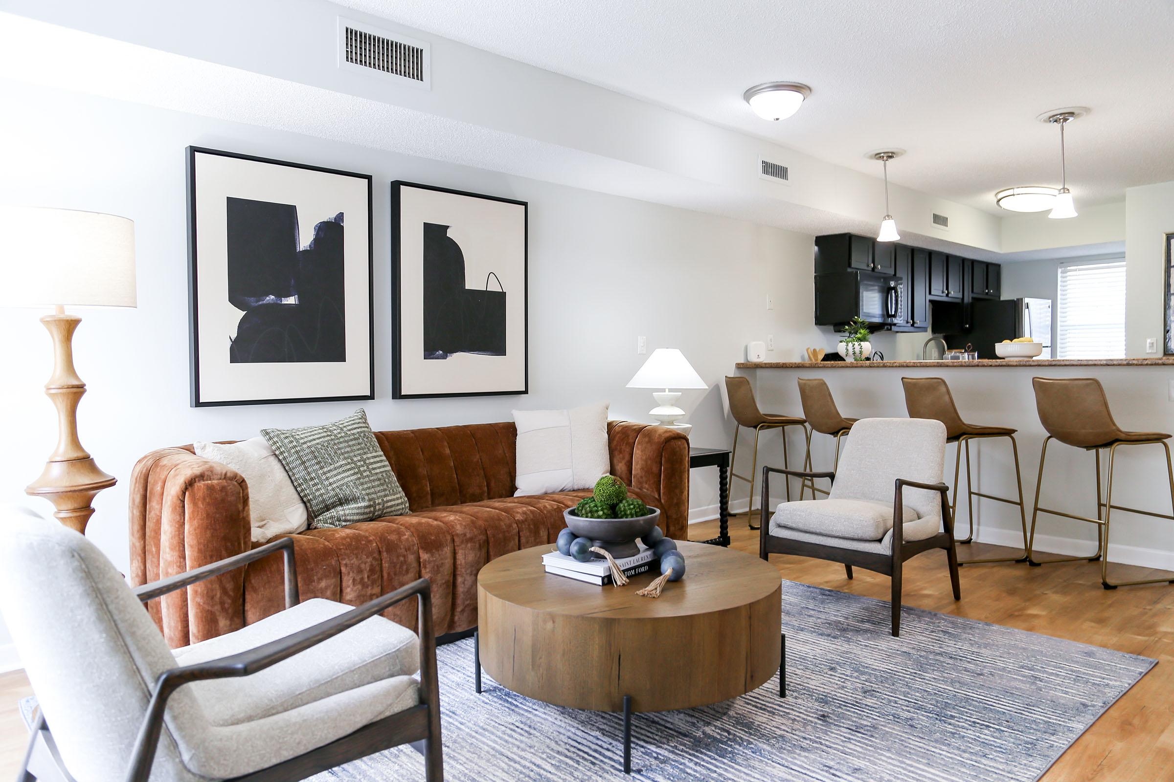 A stylish living room featuring a brown velvet sofa with decorative pillows, two framed artworks on the wall, a round wooden coffee table, and four modern bar stools at a kitchen island in the background. The space is bright and inviting with natural light and neutral decor.