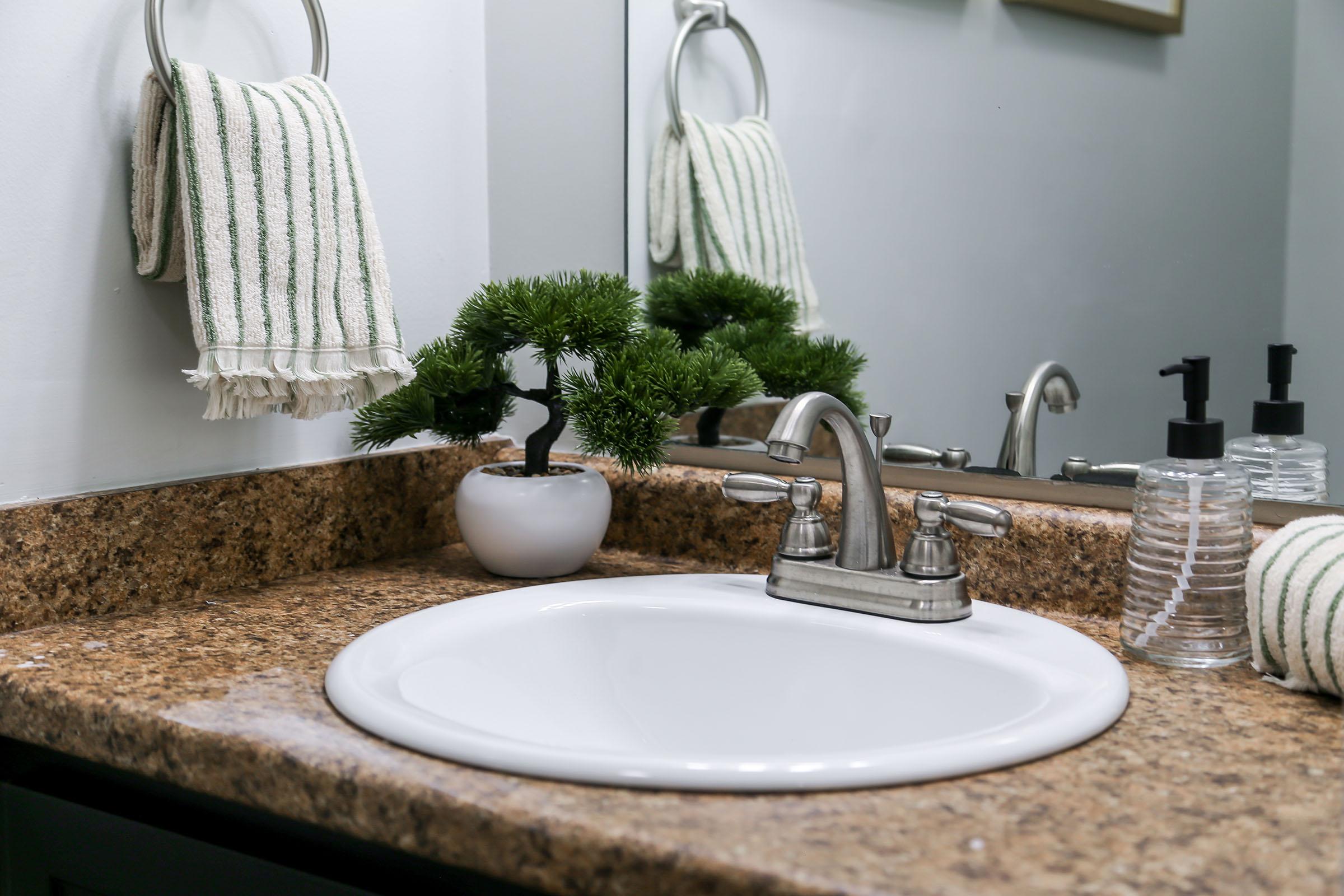 A bathroom sink with a white basin and chrome faucet, set on a speckled granite countertop. A small potted plant adds a touch of greenery, and there are two black soap dispensers and a stack of clear plastic cups nearby. A striped hand towel hangs on a ring mounted on the wall, with a mirror reflecting the scene.