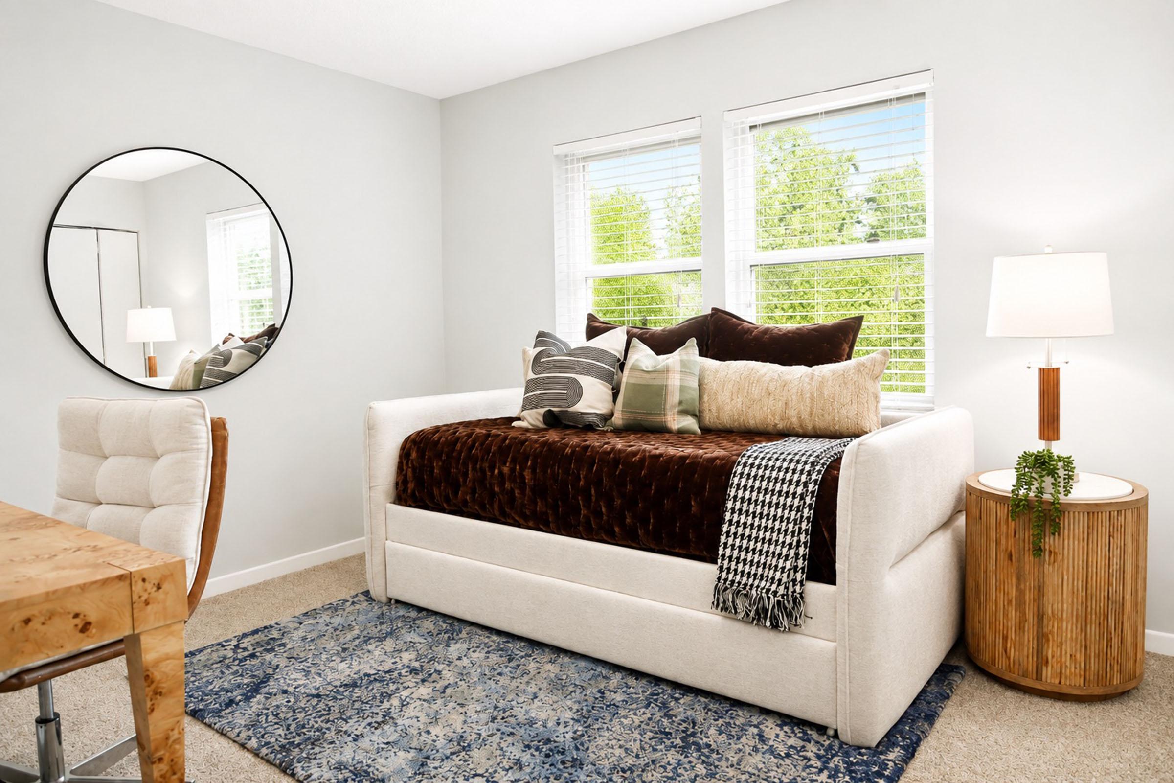 A cozy, well-lit room featuring a daybed with brown and cream-colored pillows and a textured blanket. There's a round mirror on the wall opposite the bed, a stylish wooden desk with a plush chair, and a side table with a lamp. Sunlight filters in through the windows, illuminating the soft color palette.