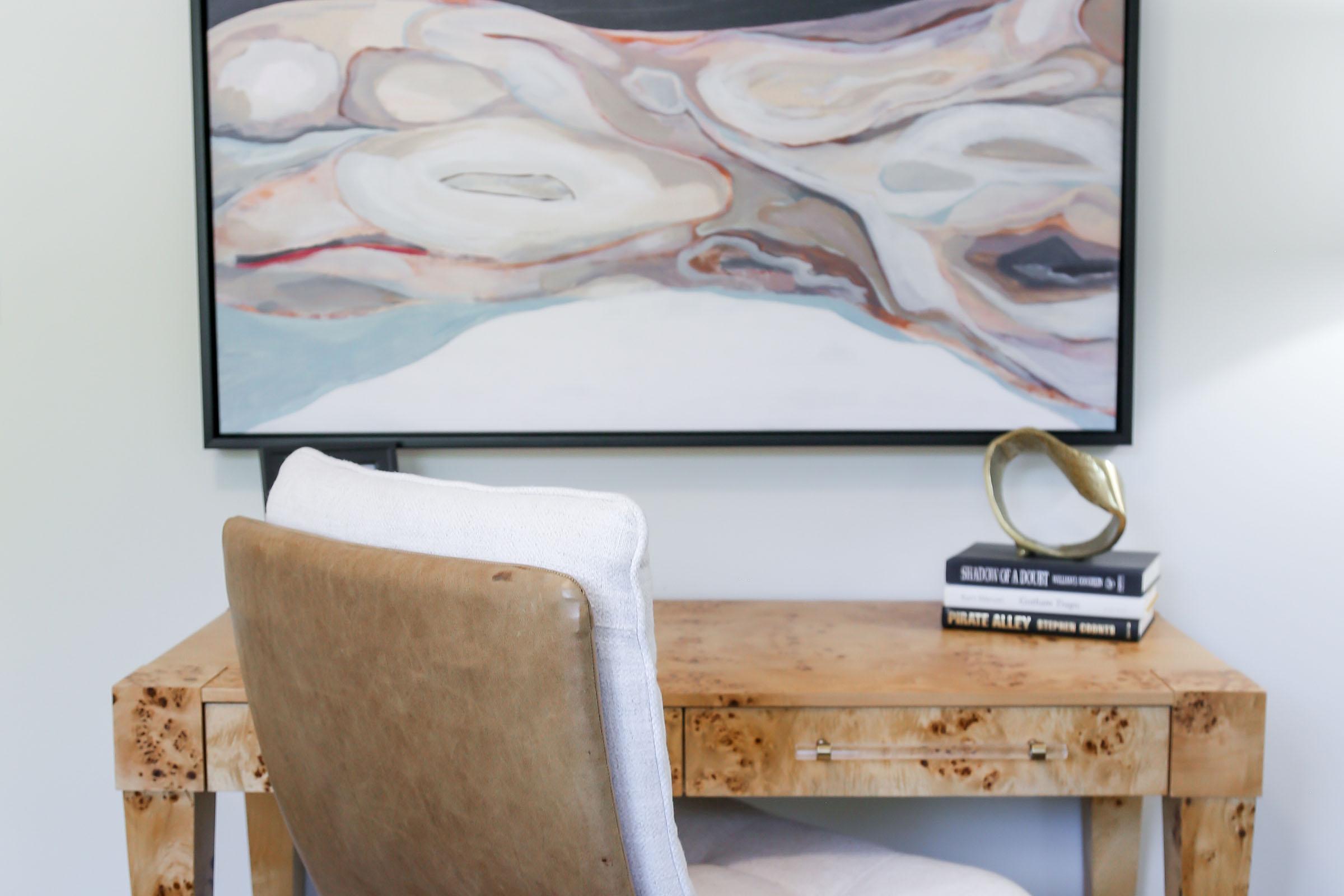 A modern workspace featuring a wooden desk with a stylish chair. On the desk, there are a few stacked books and a decorative object, with a large abstract painting hanging on the wall above it. The overall aesthetic is contemporary and minimalistic, creating a calm work environment.