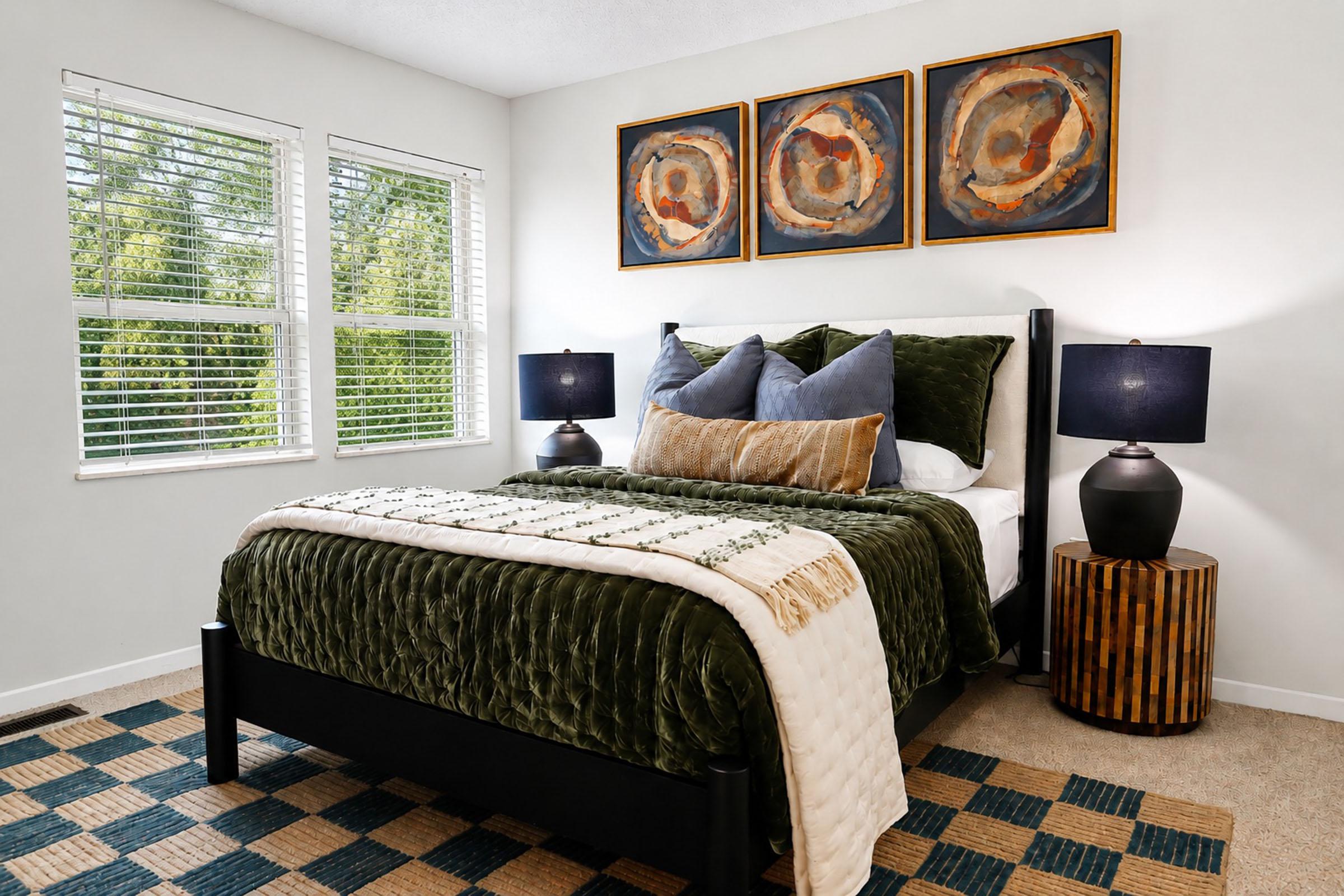 A cozy bedroom featuring a dark green bedspread, decorative pillows, and a quilt. Two lamps sit on bedside tables next to the bed. Above the bed, three abstract framed artworks adorn the wall. Natural light streams in through two large windows with white blinds, illuminating the warm, neutral-toned decor.