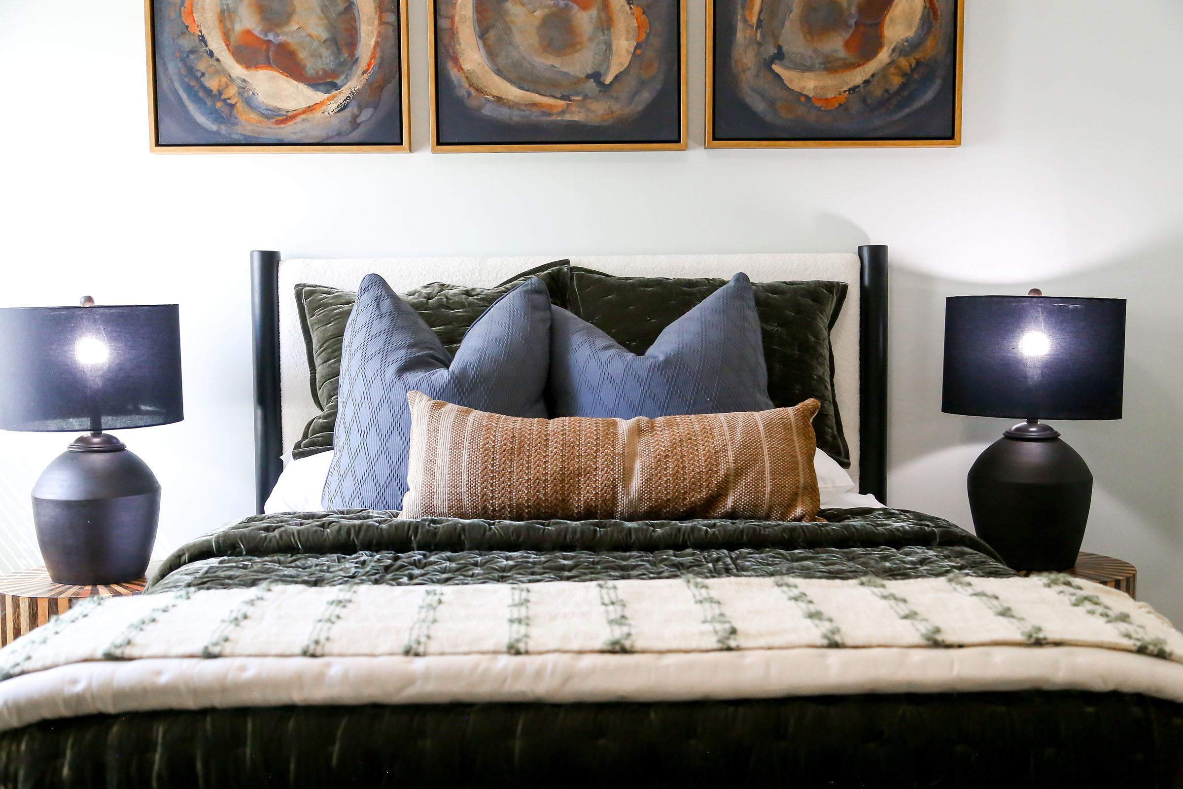 A cozy bedroom setup featuring a neatly made bed with an assortment of pillows. The bed is adorned with a dark green throw blanket and a light-colored quilt. On either side of the bed, there are black table lamps with warm light, and above the bed, three abstract framed artworks in gold and earthy tones.