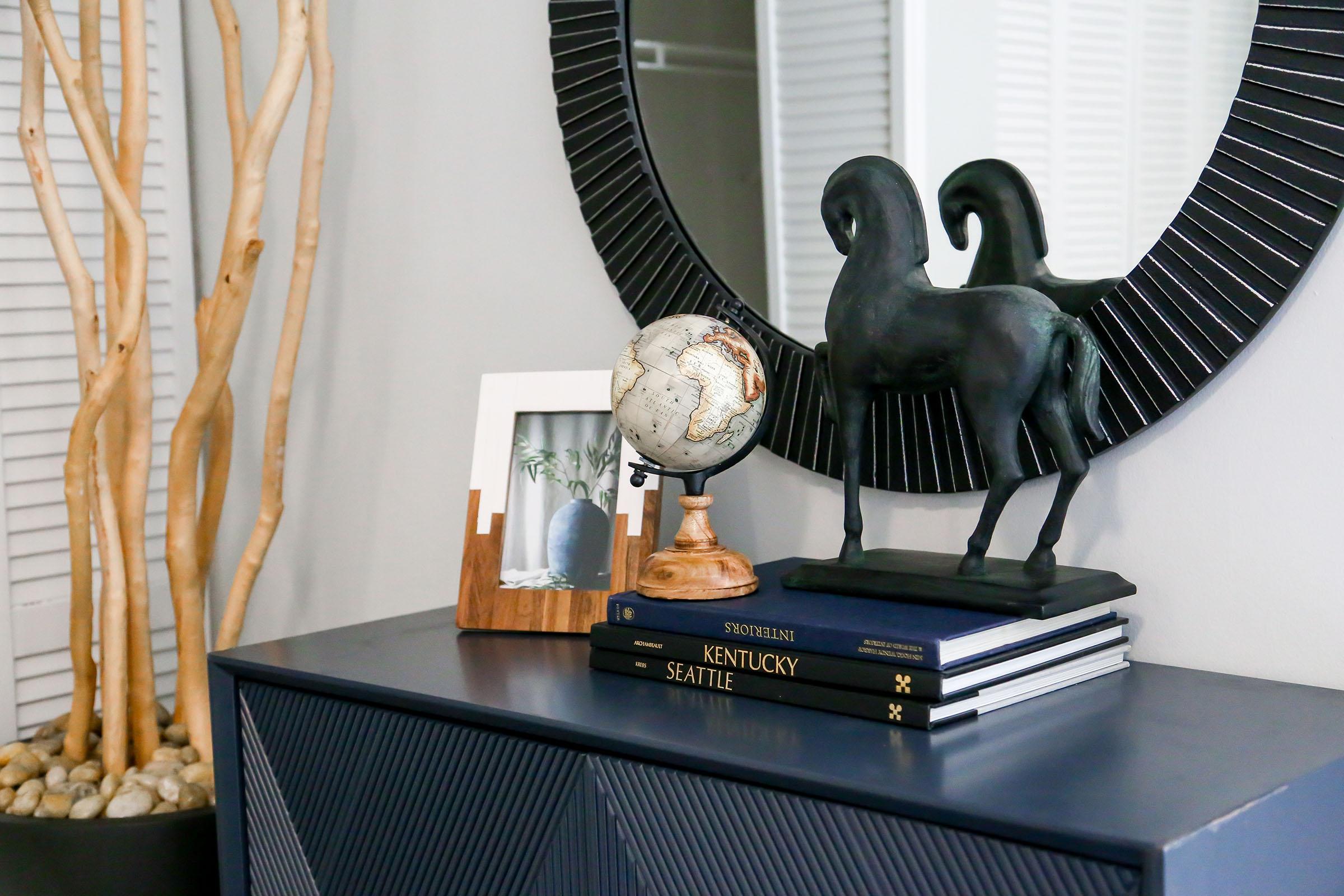 A stylish home decor scene featuring a round mirror, a decorative globe on a wooden stand, a black sculpture of two rams, and stacked books titled "Kentucky" and "Seattle." Tall, slender decorative branches and a framed photo are also present, arranged on a dark blue cabinet.