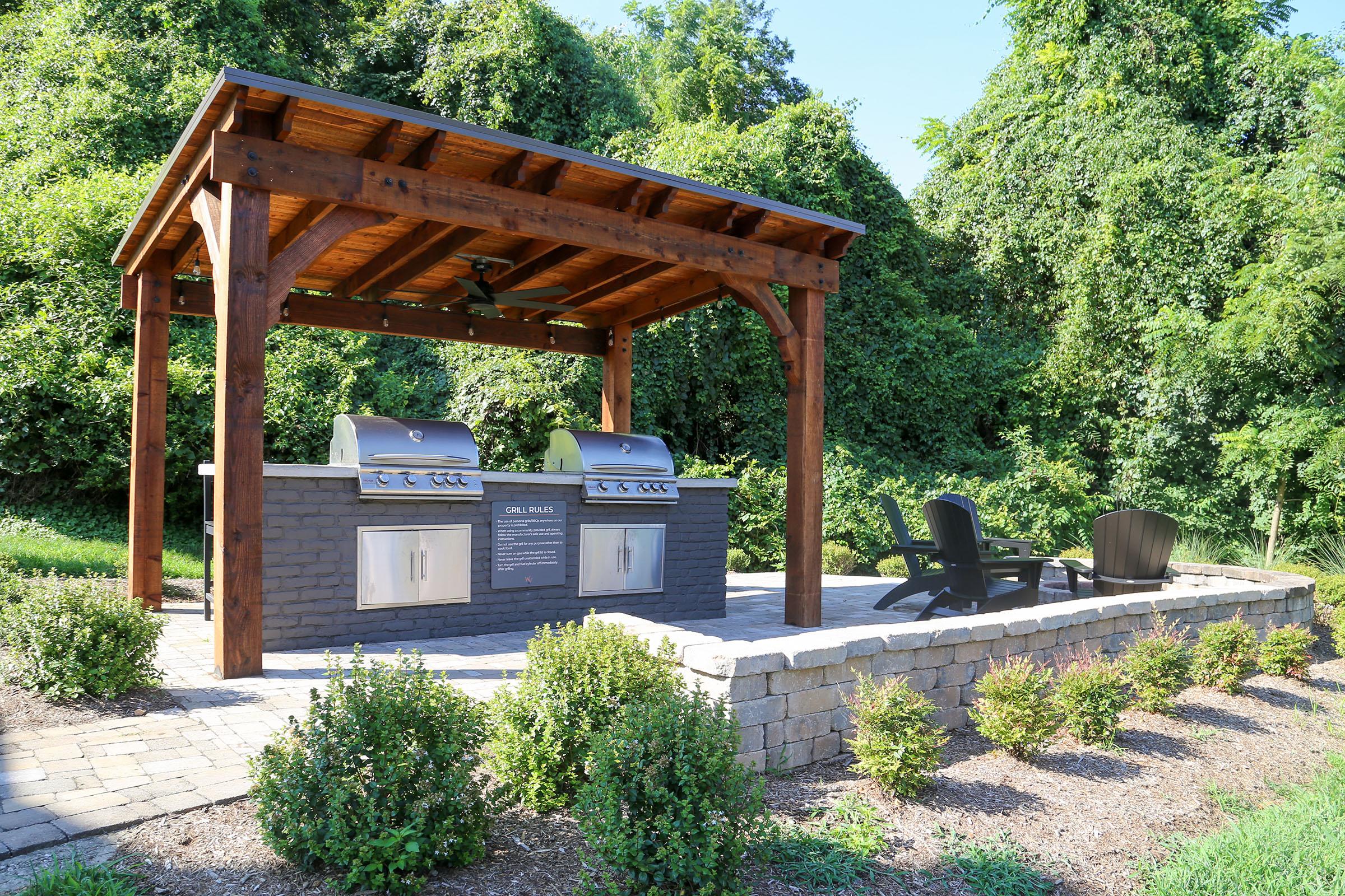 Outdoor grilling area featuring a wooden pergola, two stainless steel grills, and a stone base. Surrounding the space are neatly trimmed shrubs and a paved patio, complemented by two black Adirondack chairs for seating. Lush greenery serves as a backdrop, creating a serene outdoor atmosphere.