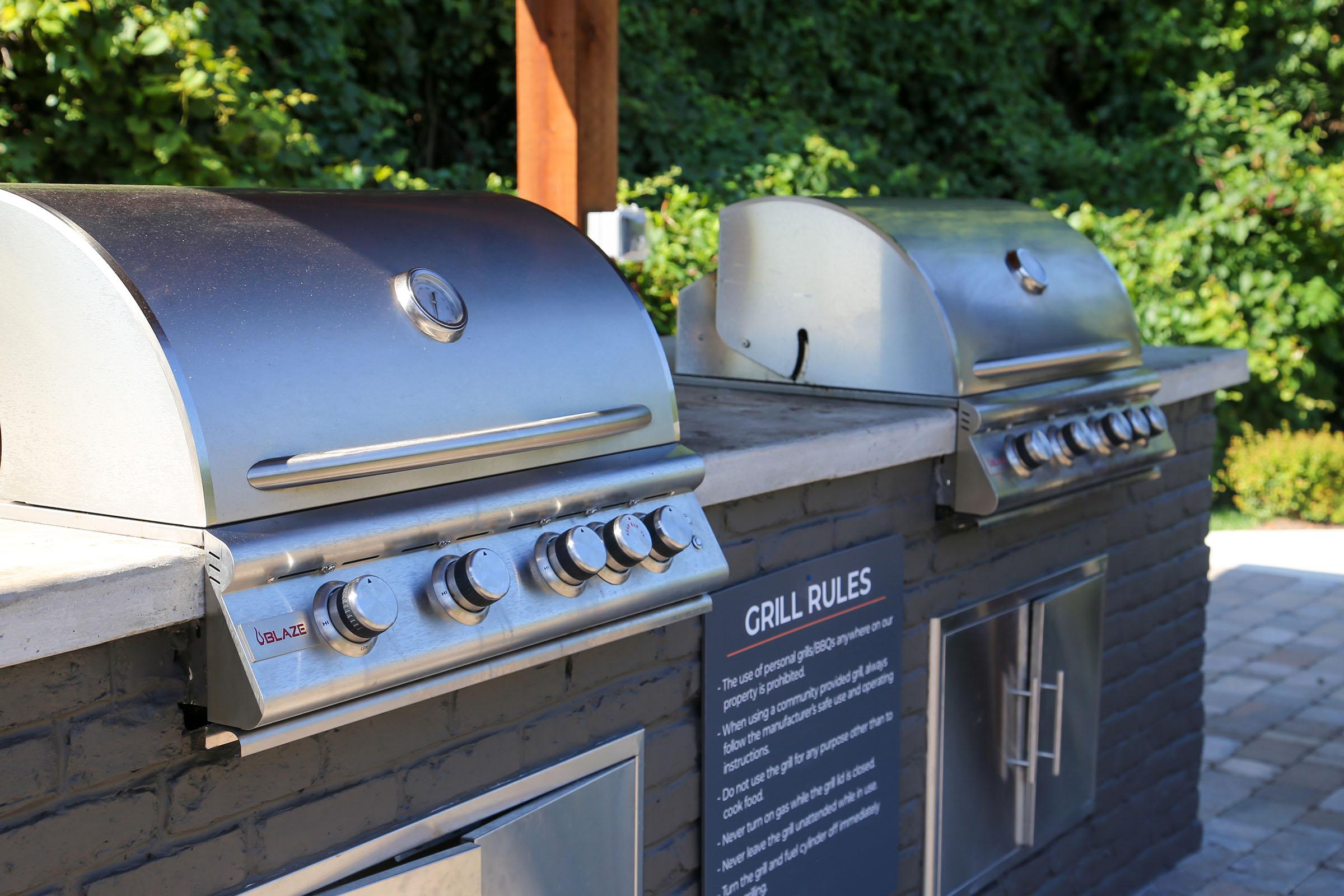 Two stainless steel gas grills on a stone countertop, with a sign displaying "Grill Rules" nearby. The background features lush greenery, suggesting an outdoor cooking area designed for gatherings and barbecues. The grills have multiple control knobs for temperature adjustment.