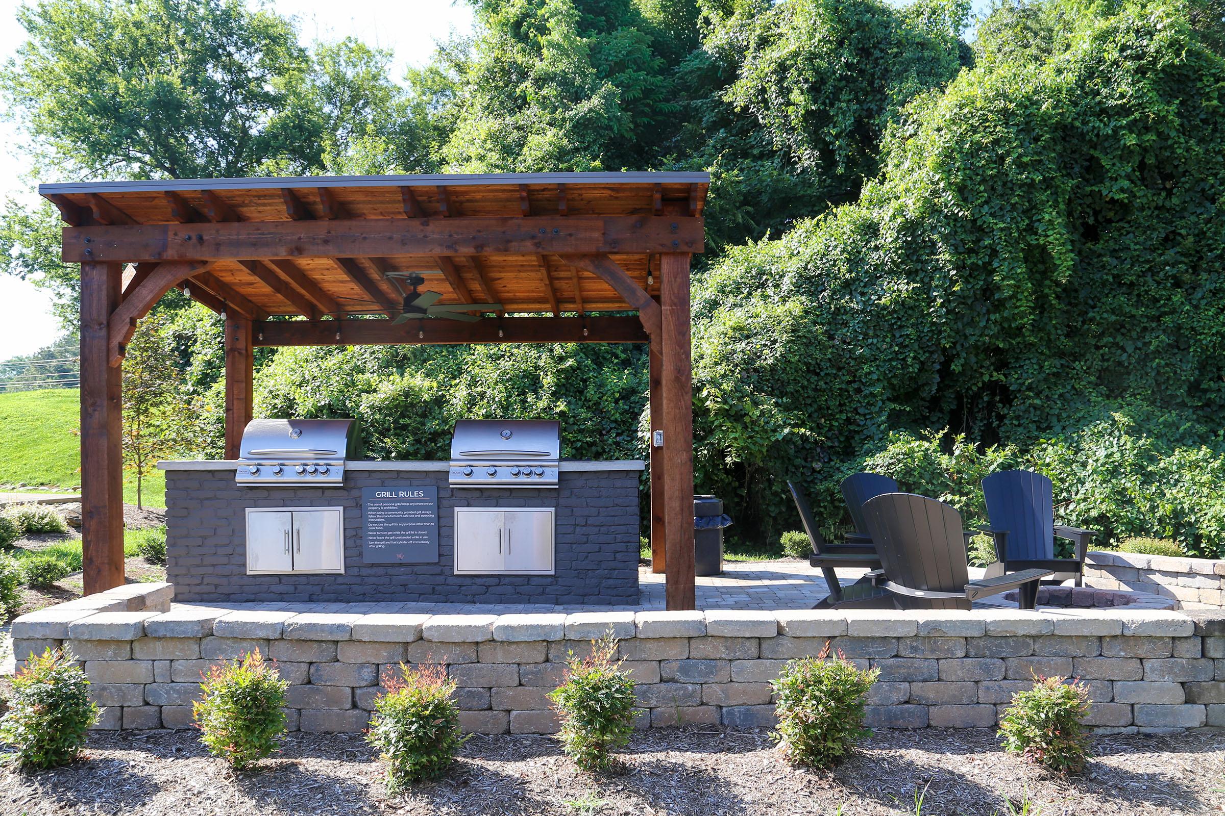 An outdoor grilling area featuring two stainless steel grills under a wooden pergola. The space includes a stone countertop, seating area with dark Adirondack chairs, and a landscaped garden with low shrubs along the front. Lush greenery is visible in the background.