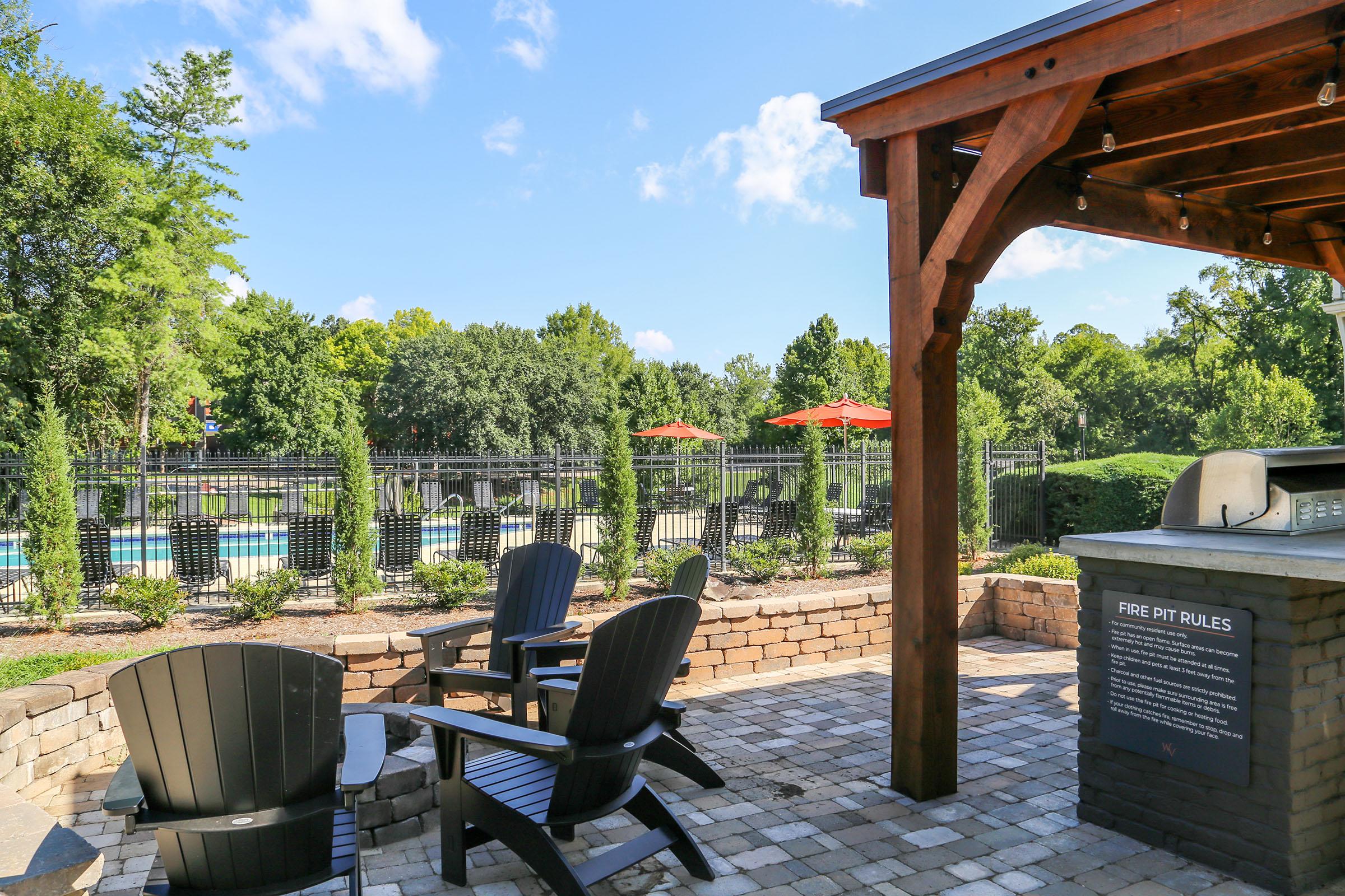A cozy outdoor area featuring a fire pit surrounded by dark wooden chairs. In the background, there is a swimming pool enclosed by a black fence, with vibrant red umbrellas providing shade. Lush trees and landscaping enhance the inviting atmosphere under a clear blue sky.