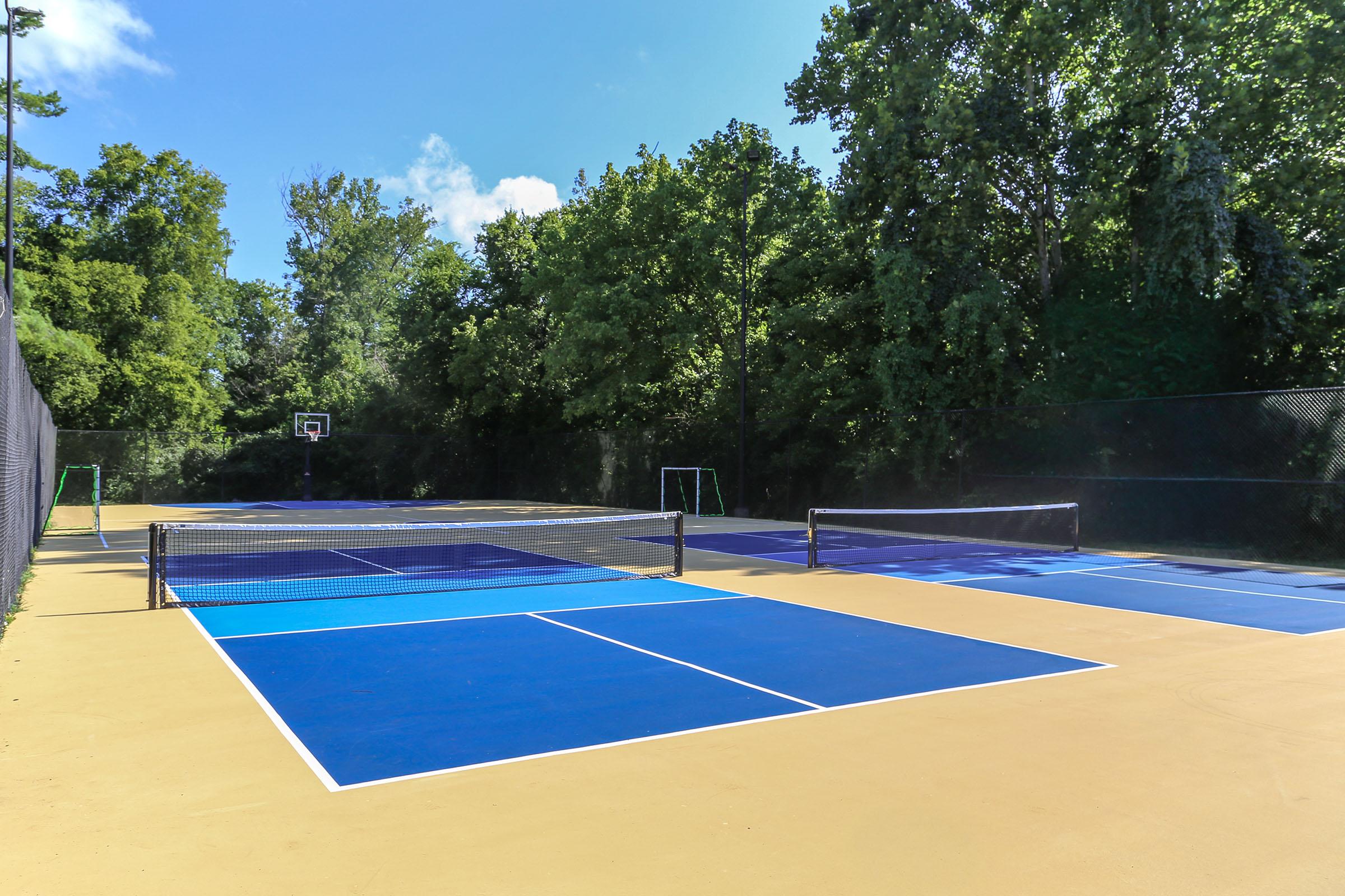A view of outdoor sports courts, primarily featuring blue tennis courts with a yellow surface. In the background, there is a basketball hoop and greenery surrounding the area. The scene is bright and sunny, conveying a recreational atmosphere.