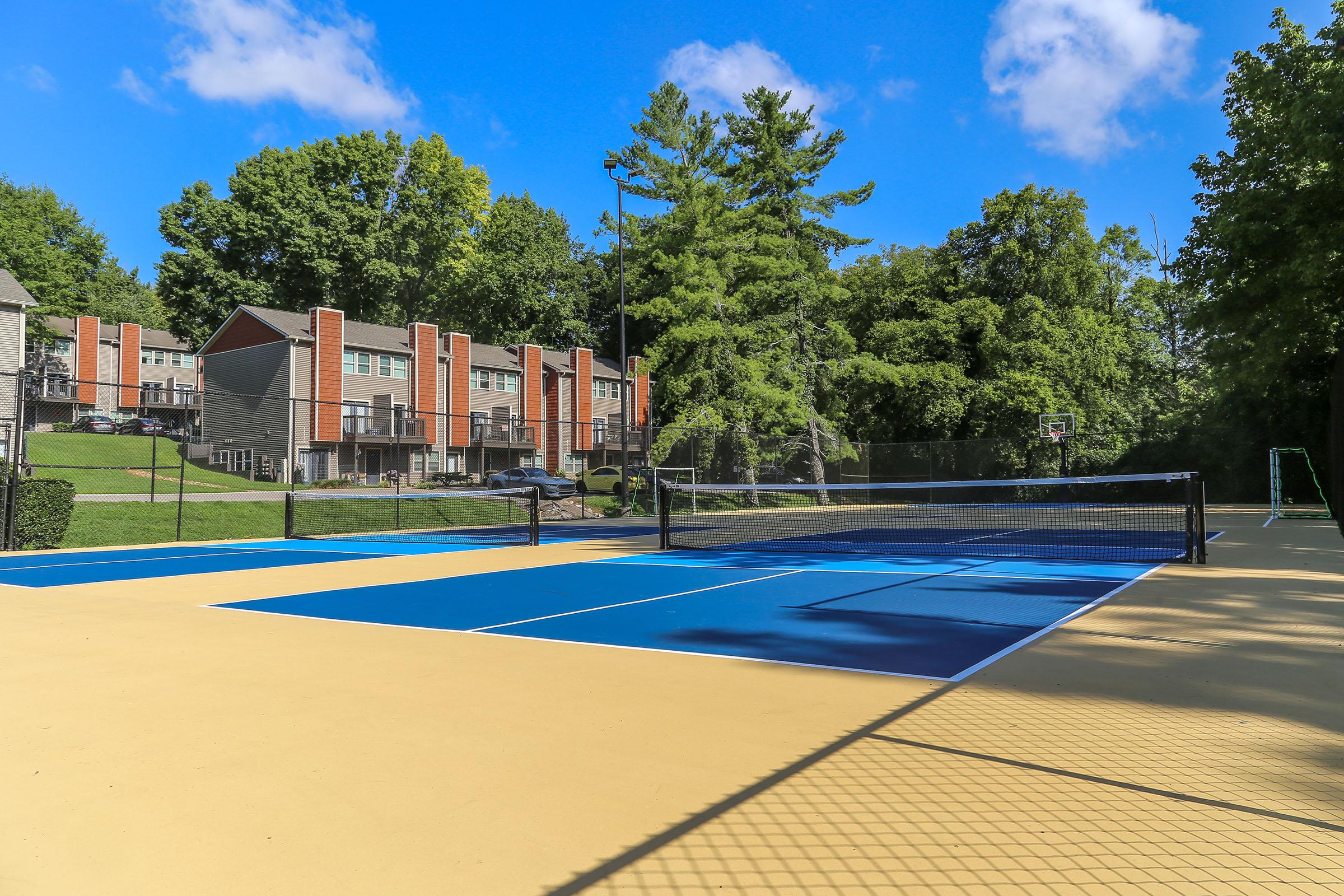 A bright outdoor sports court featuring blue tennis courts with a net, surrounded by lush green trees and residential buildings in the background under a clear blue sky. A basketball hoop is visible on the side, completing the recreational setting.