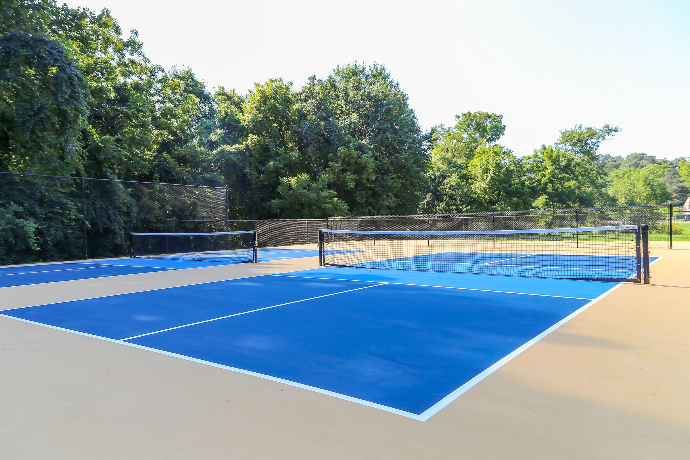A tennis court featuring blue playing surfaces and black nets, surrounded by green trees and a clear sky. The court is empty, highlighting the smooth asphalt and marked lines. The scene conveys a peaceful outdoor setting ideal for tennis play.
