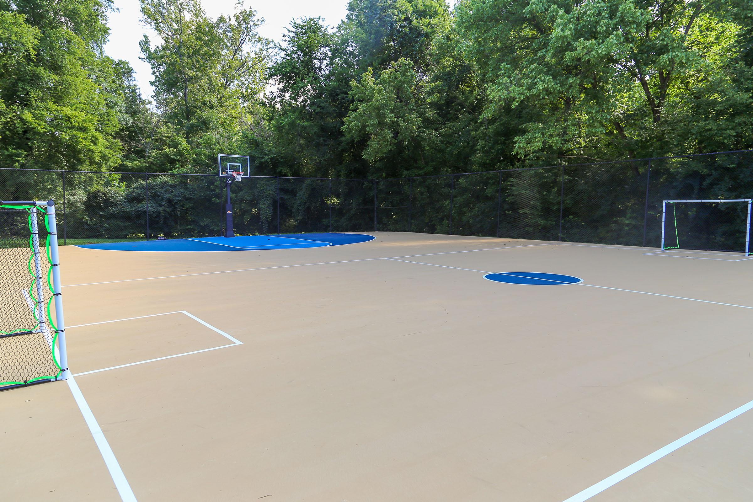 A multi-use sports court surrounded by trees, featuring a basketball hoop at one end and two soccer goals at each side. The court has a smooth, beige surface with blue circular markings and is enclosed by a fence, providing a versatile space for outdoor activities.