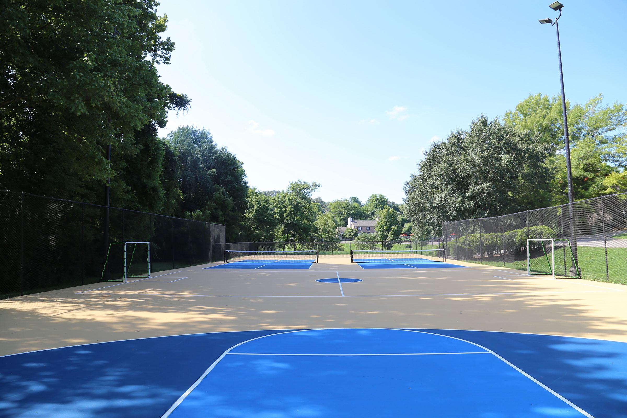 A view of a well-maintained outdoor basketball court with vibrant blue and yellow surfaces, surrounded by trees. There are two small soccer goals at either end of the court, and a clear blue sky overhead. The area is quiet, displaying a peaceful and inviting atmosphere for sports and recreation.