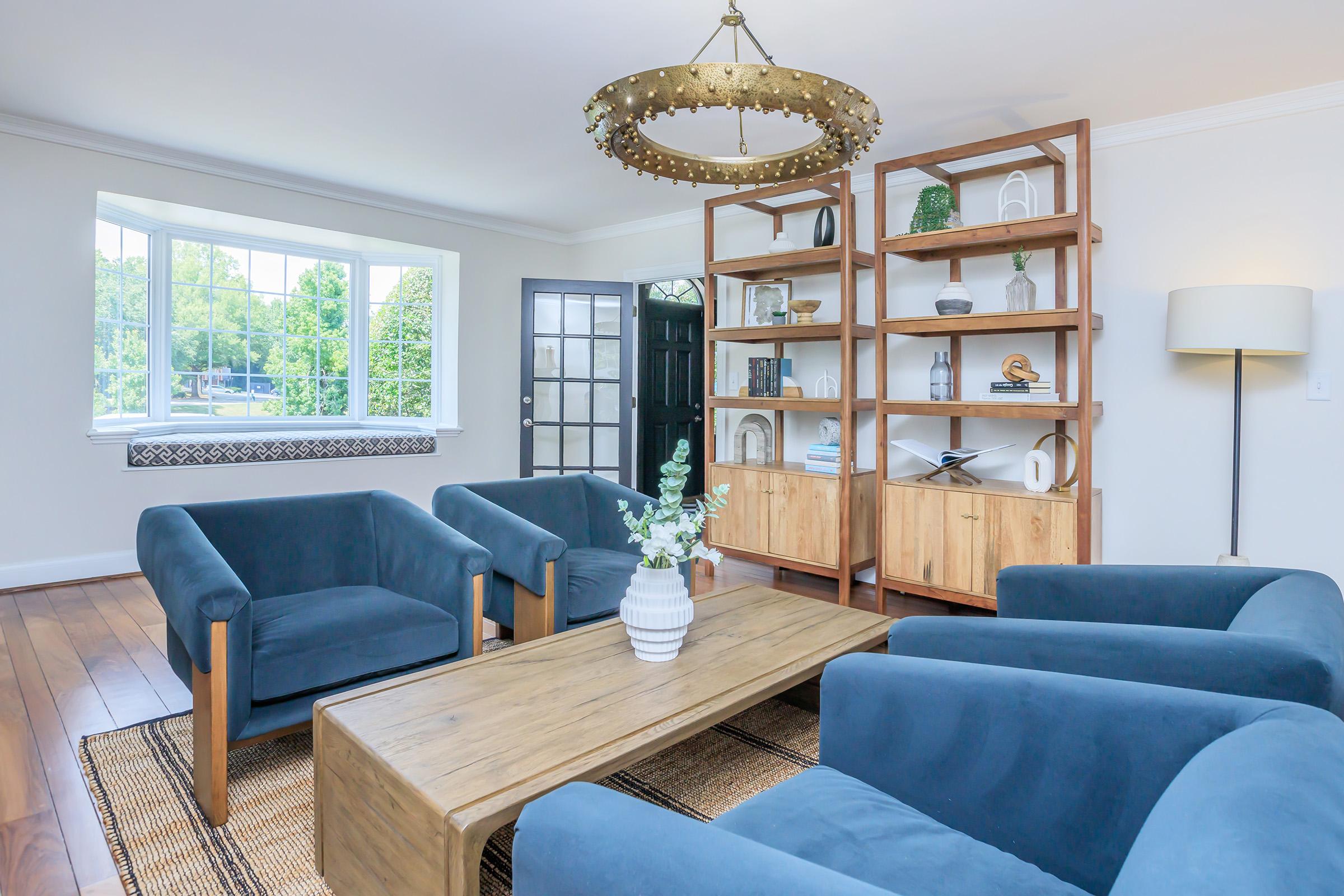 A cozy living room featuring two blue armchairs and a wooden coffee table. A stylish bookshelf filled with decorative items stands against the wall. Natural light streams in through a large window, illuminating the space, while a floor lamp adds warmth. The flooring is wooden, enhancing the room's inviting atmosphere.