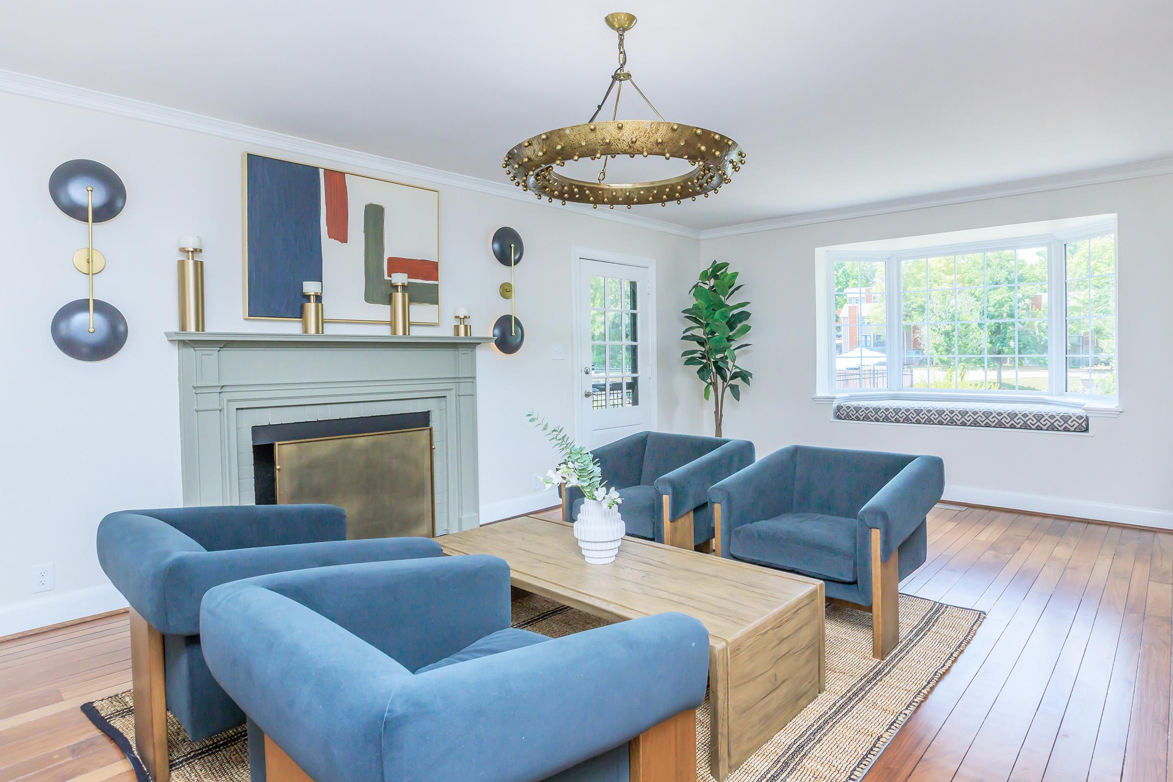A modern living room featuring a light-colored wall and hardwood flooring. It includes a stylish fireplace with a decorative mantel, a wooden coffee table, and four blue upholstered chairs arranged around it. A large window offers natural light, and a potted plant adds a touch of greenery.