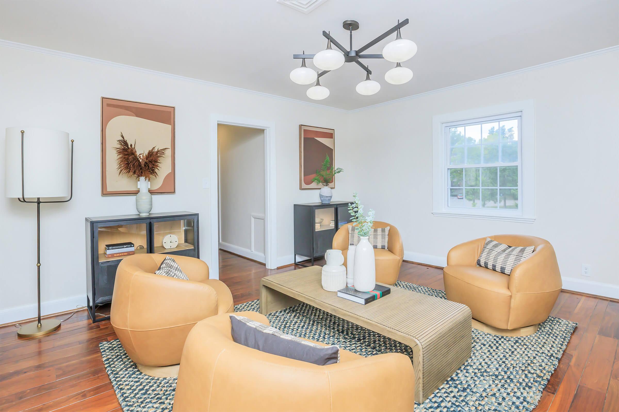 A stylish living room featuring three tan armchairs arranged around a modern coffee table. Light-colored walls and hardwood flooring complement the decor, which includes framed artwork on the walls and a potted plant on the table. A unique light fixture hangs from the ceiling, adding a contemporary touch.