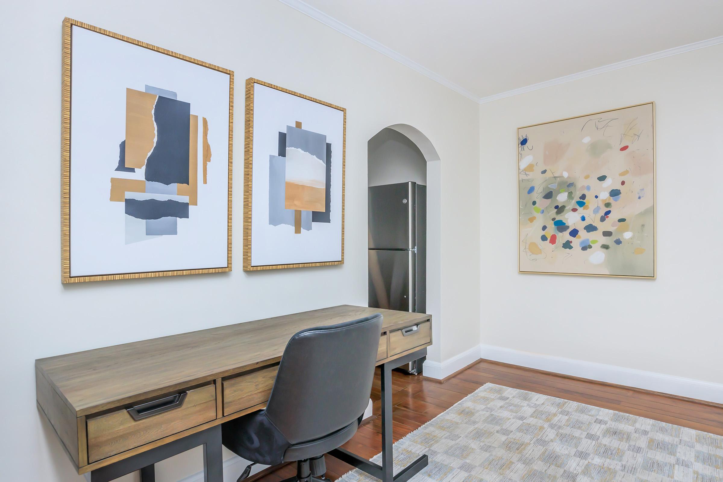 A minimalist home office scene featuring a wooden desk with a black chair, two abstract wall art pieces in gold frames, and a light-colored rug on the floor. A modern fridge is visible through an archway against a neutral wall, creating a clean and inviting atmosphere.