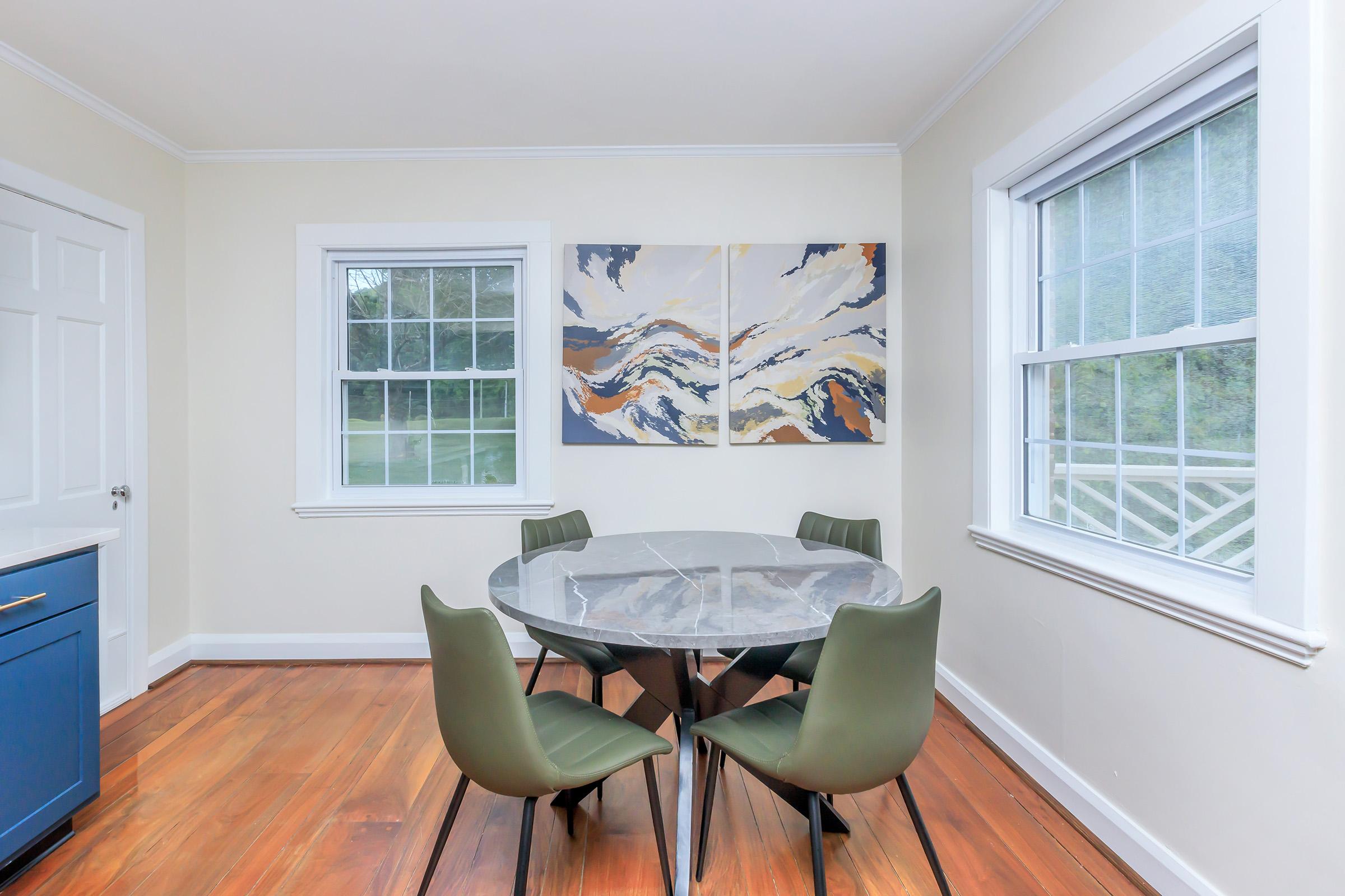 A modern dining area featuring a round marble table surrounded by green chairs. Two large windows provide natural light, and an abstract painting in warm colors hangs on the wall, complementing the contemporary style. The room has light walls and wooden flooring.