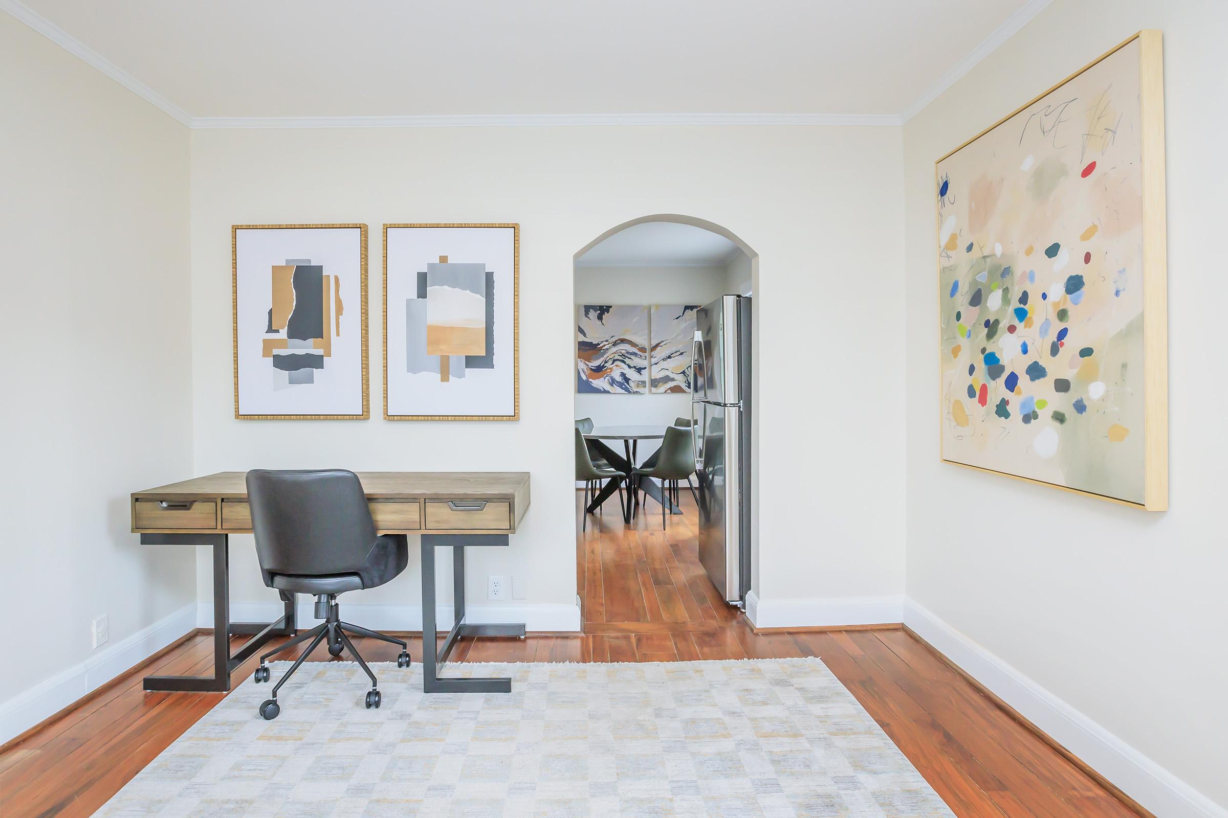 A modern home office featuring a wooden desk with a black chair, two framed abstract art pieces on the wall, and a large colorful painting. An archway leads to a kitchen area with a silver refrigerator and additional seating. The flooring is wooden, and a neutral-colored rug lies beneath the desk.