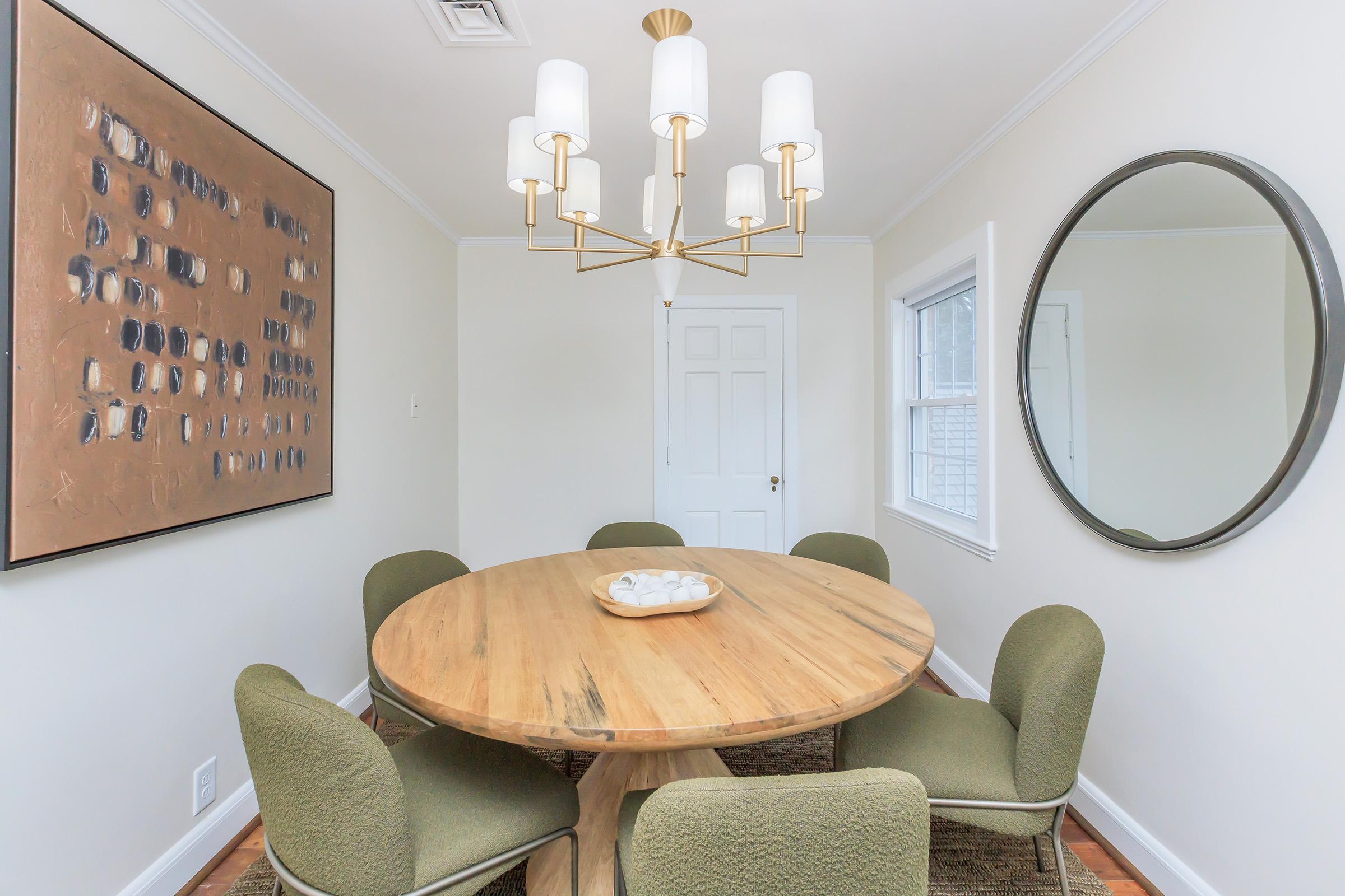 A modern dining room featuring a round wooden table surrounded by green upholstered chairs. Above hangs a stylish chandelier with multiple white shades. A circular mirror adorns the wall, complemented by a framed artwork with abstract patterns. Natural light filters through a window.