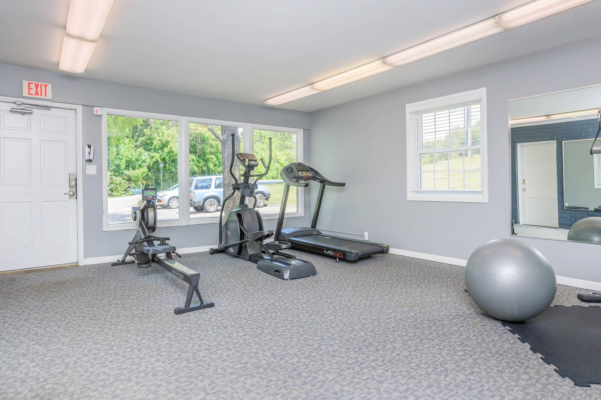 A small gym or fitness room featuring exercise equipment such as a treadmill, elliptical machine, rowing machine, and an exercise ball. There are large windows letting in natural light, gray walls, and a mirror on one wall, creating a spacious atmosphere.