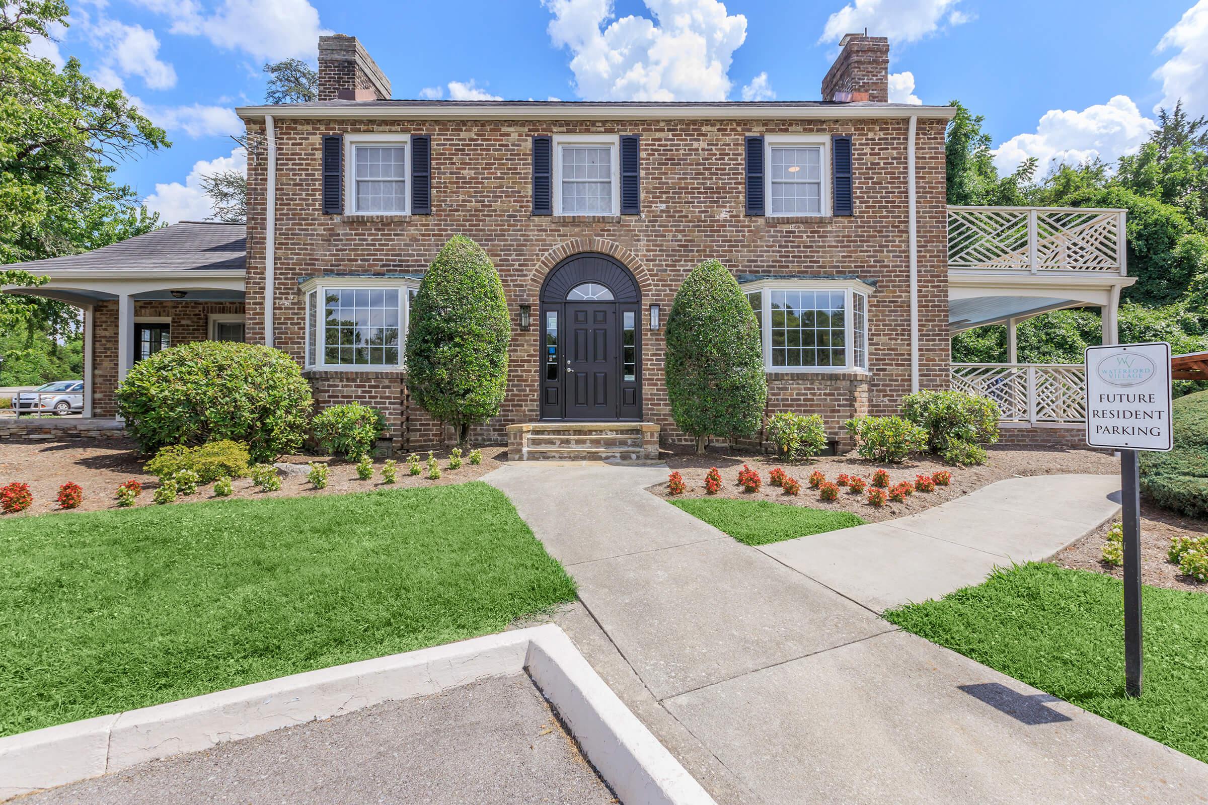 A two-story brick building with a black front door, symmetrical windows, and neatly trimmed shrubs. A sign indicating "Future Resident Parking" is visible in the foreground, with a landscaped path leading to the entrance and flower beds along the walkways. Blue skies with scattered clouds are in the background.
